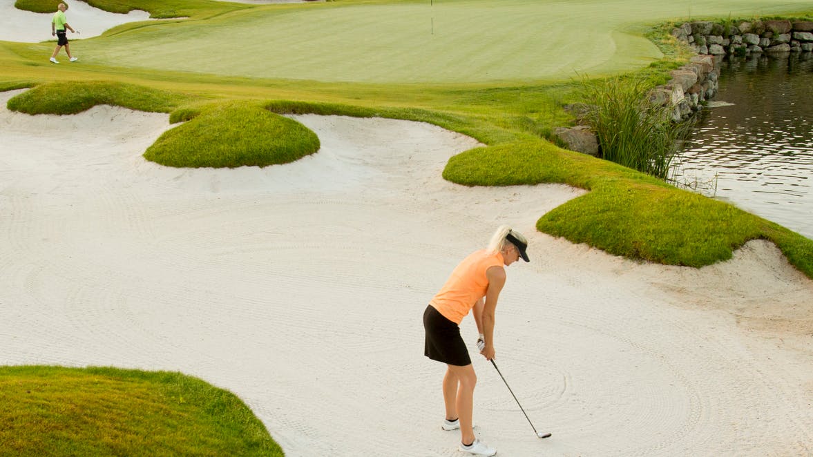 A woman in an orange shirt and black skirt prepares to hit a shot out of a sand trap