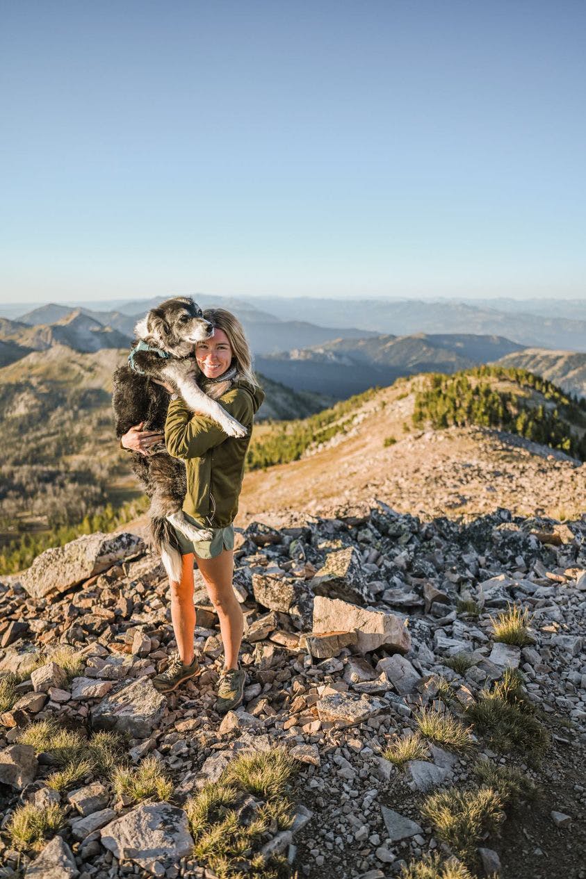 A woman holding her dog on a mountain top. There is a rocky/dirt trail behind her. 