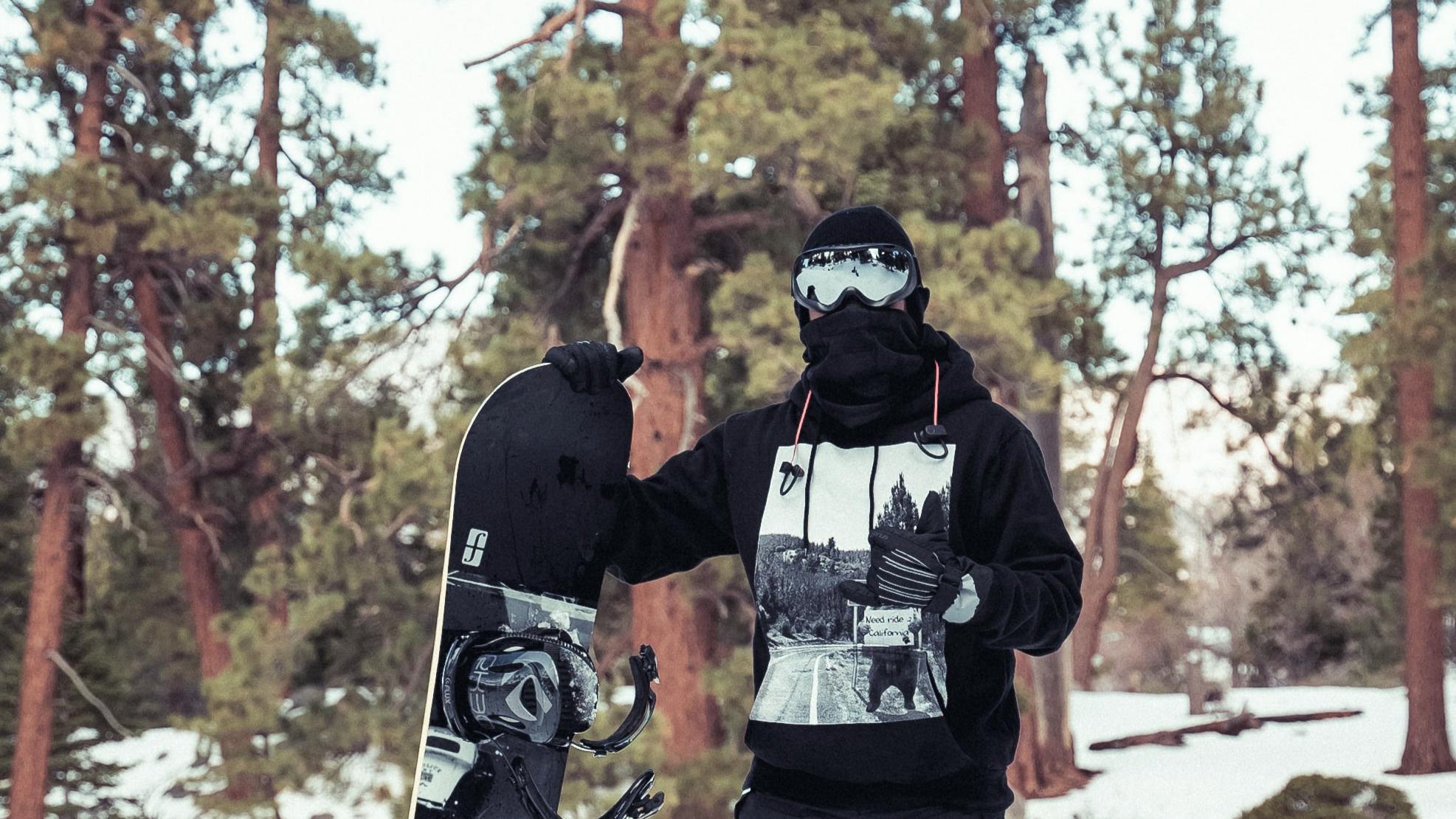 A snowboarder standing in his snowboard gear with his snowboard. There are snowy trees behind him. 