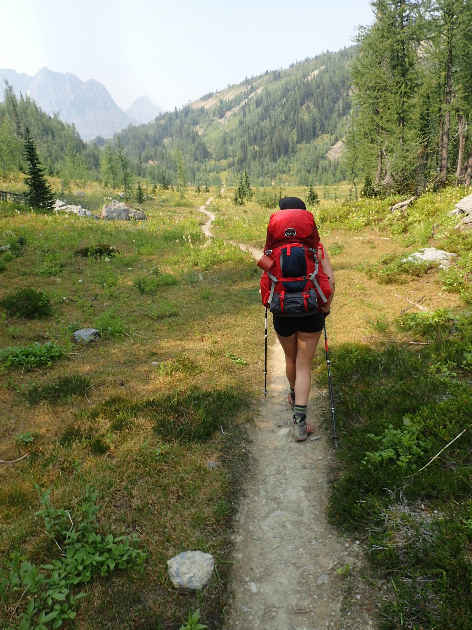 A woman with a red Osprey Ariel 65 Backpack walks down a trail. There are mountains in the background.