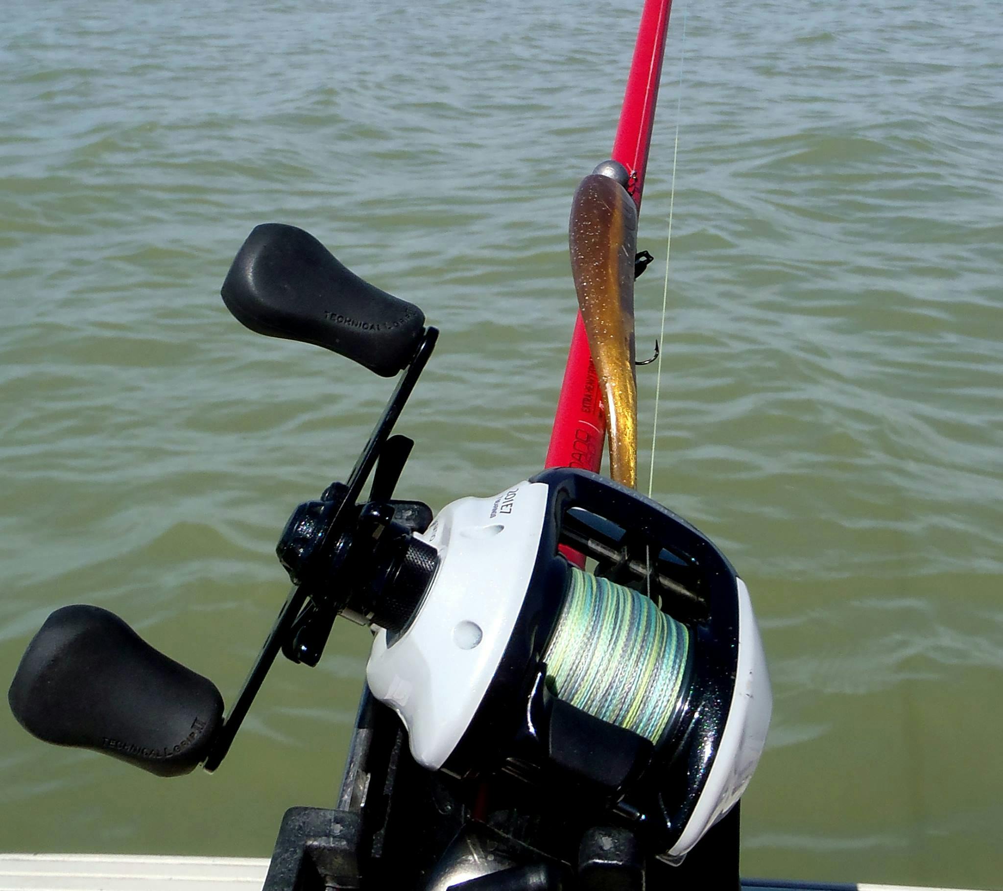 A red fishing rod with braided line on the reel is held above greenish water. 