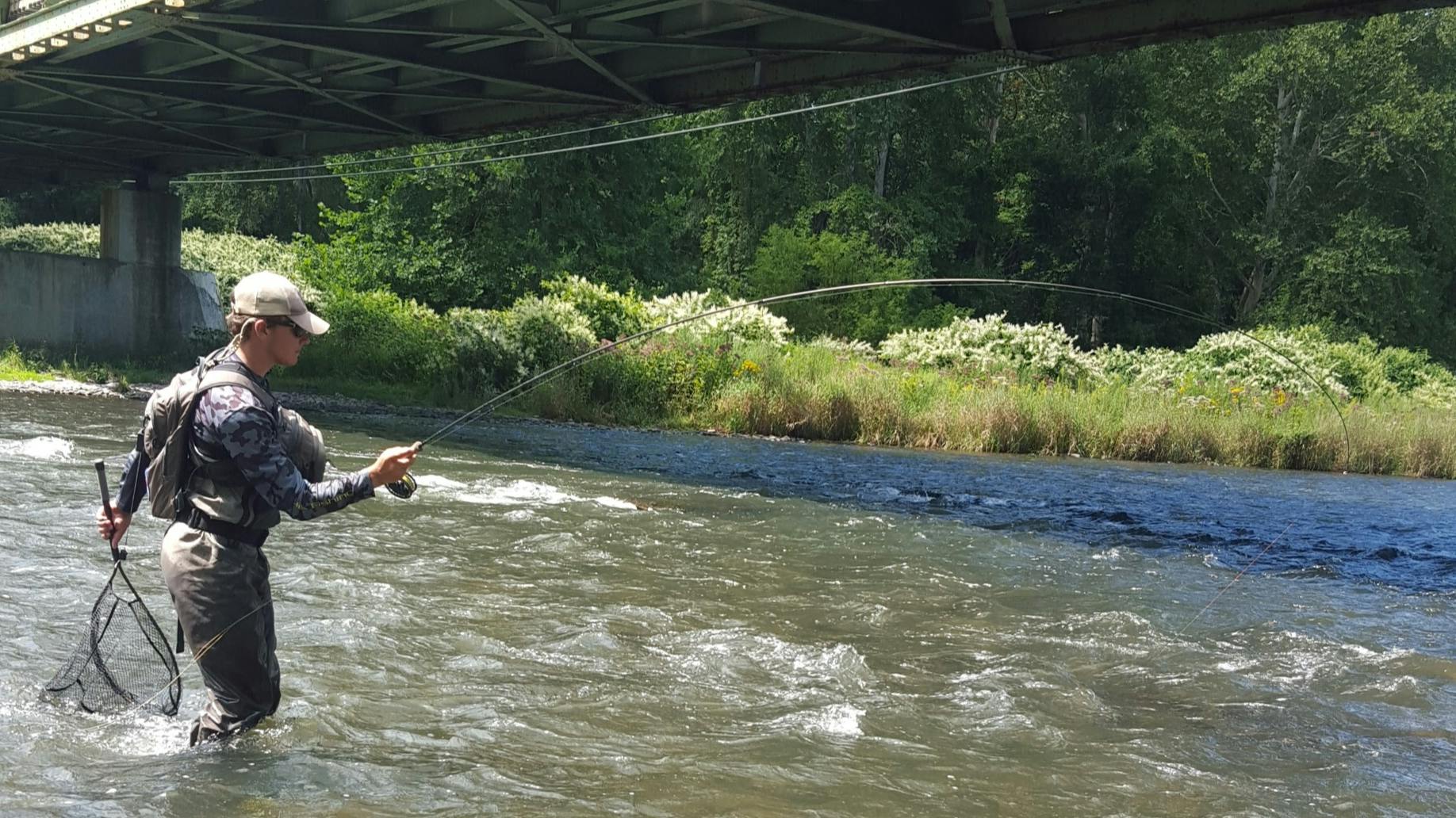 A man stands in a river under a bridge holding a fly fishing rod in one hand and a net in another