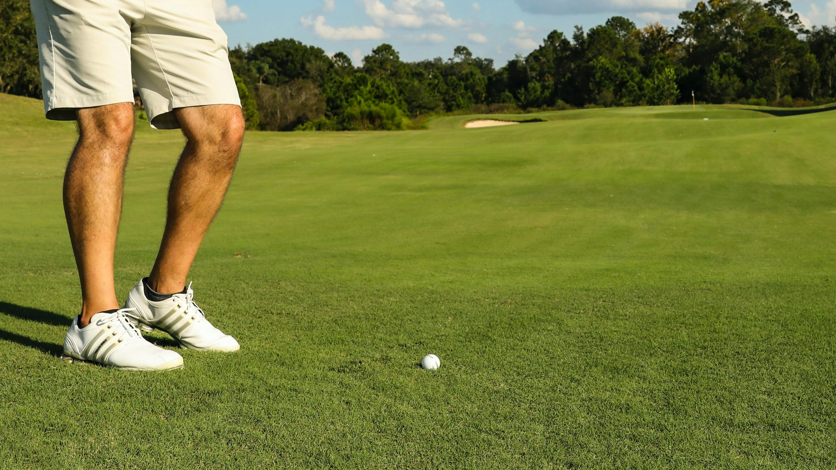 Close up of a golfers feet as he prepares to hit a golf ball. He is wearing spiked golf shoes. 