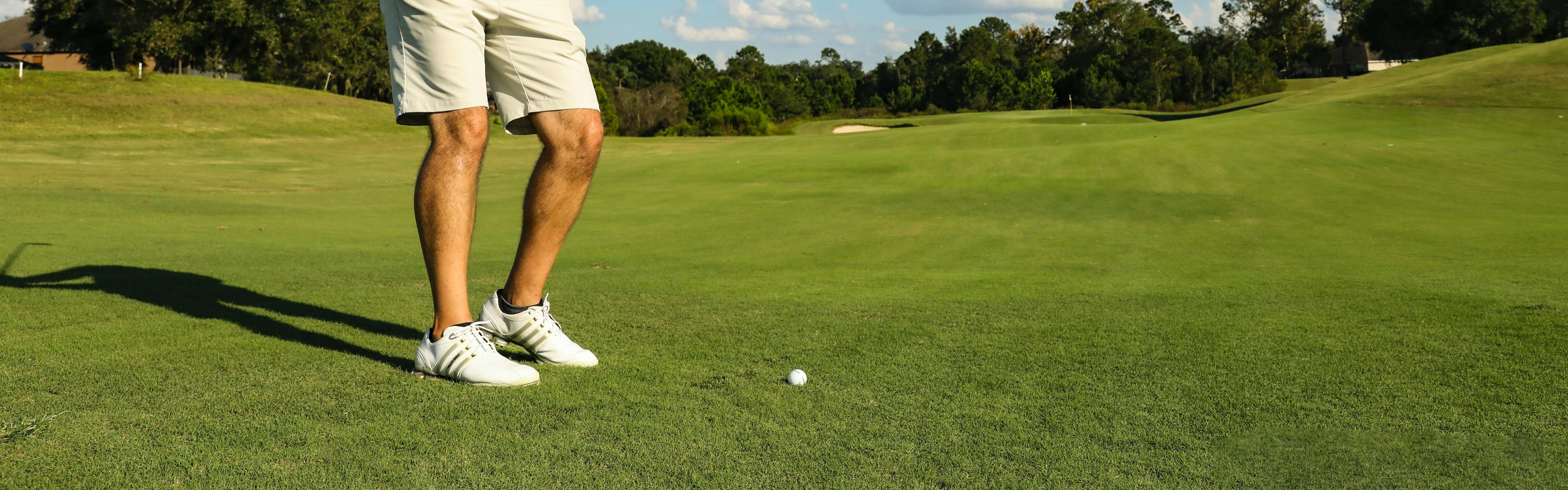 Close up of a golfers feet as he prepares to hit a golf ball. He is wearing spiked golf shoes. 