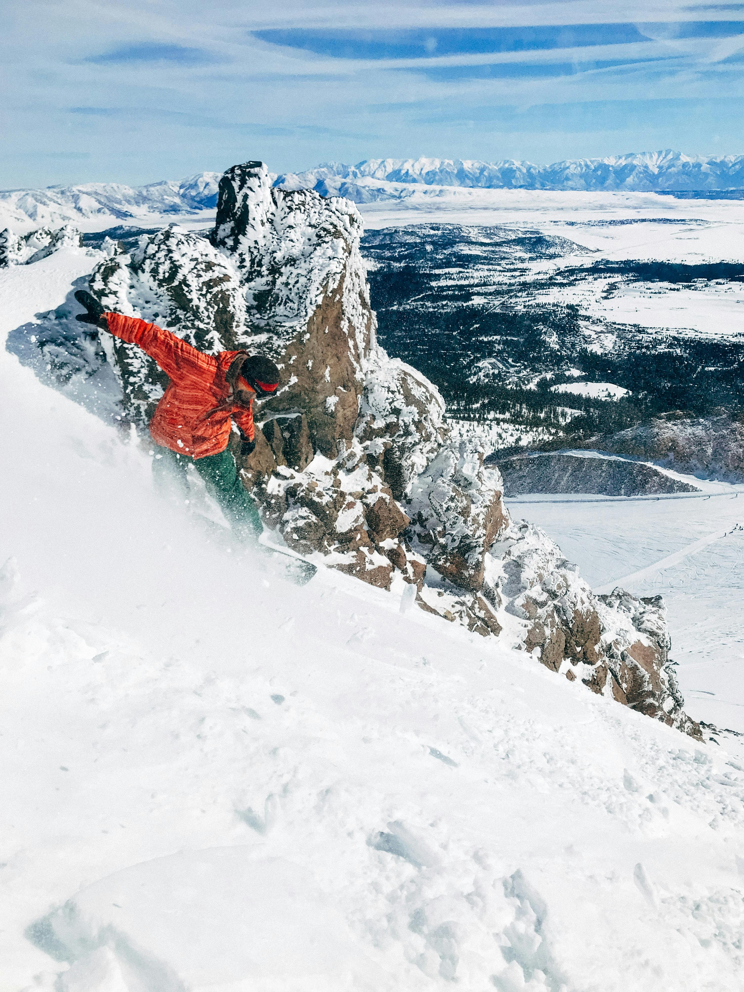 A snowboarder in an orange jacket makes their way down a steep slope with rocks and mountains in the background