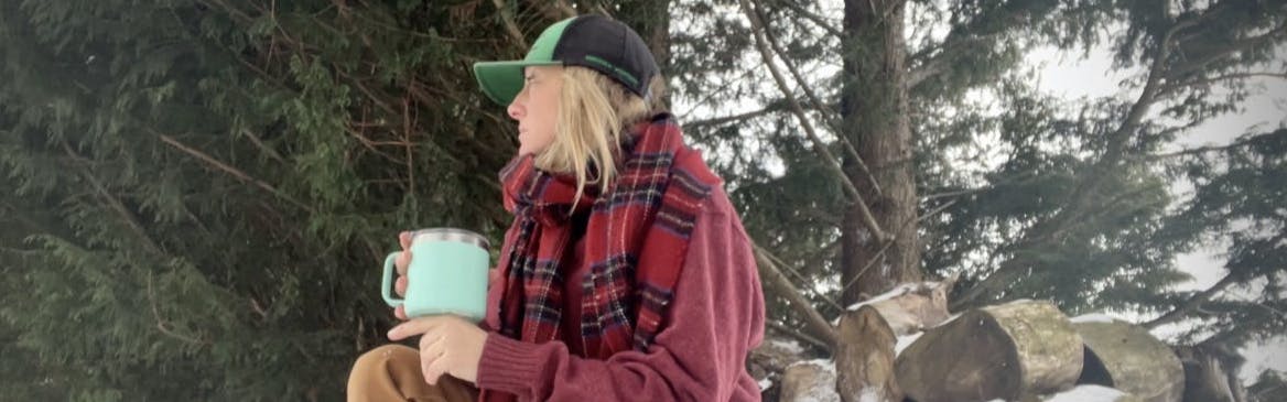 The author crouches in the snow with her Yeti mug. She wears a red sweater and scarf and a baseball cap. 