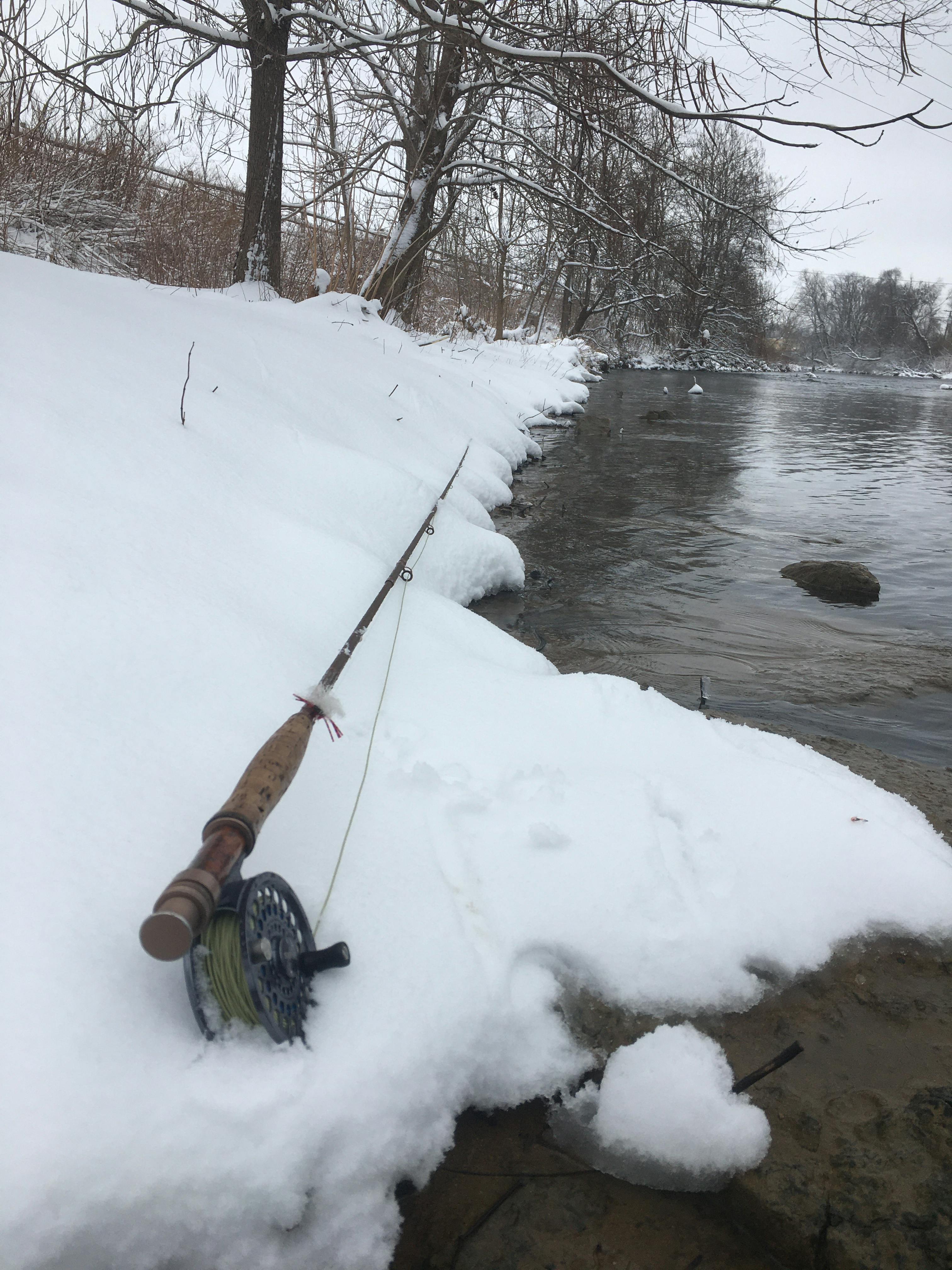 An Orvis fishing rod lies on a snowy riverbank next to the water. 
