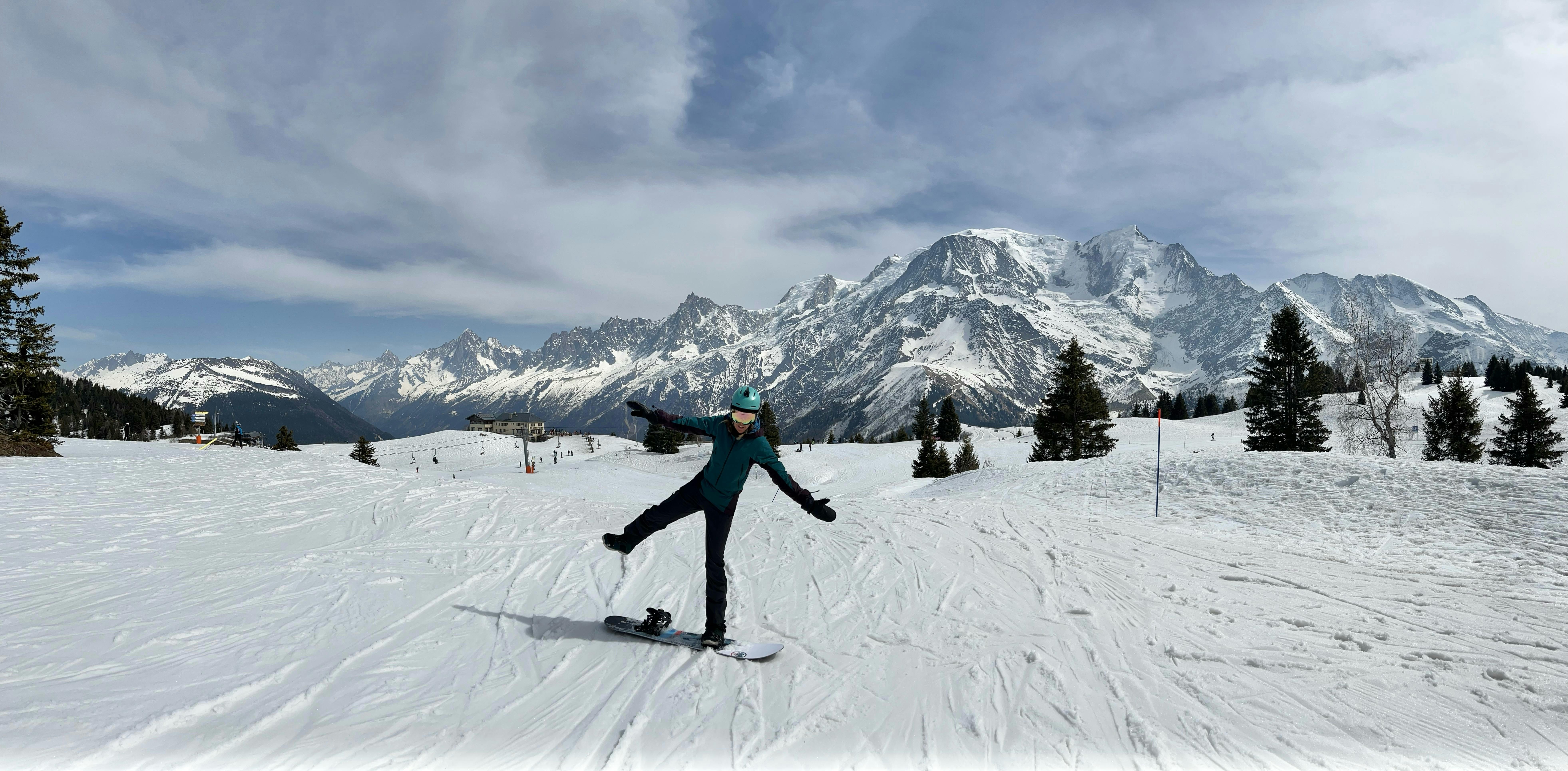 A woman stands on the Never Summer Harpoon snowboard. There are snowy mountains in the background. 