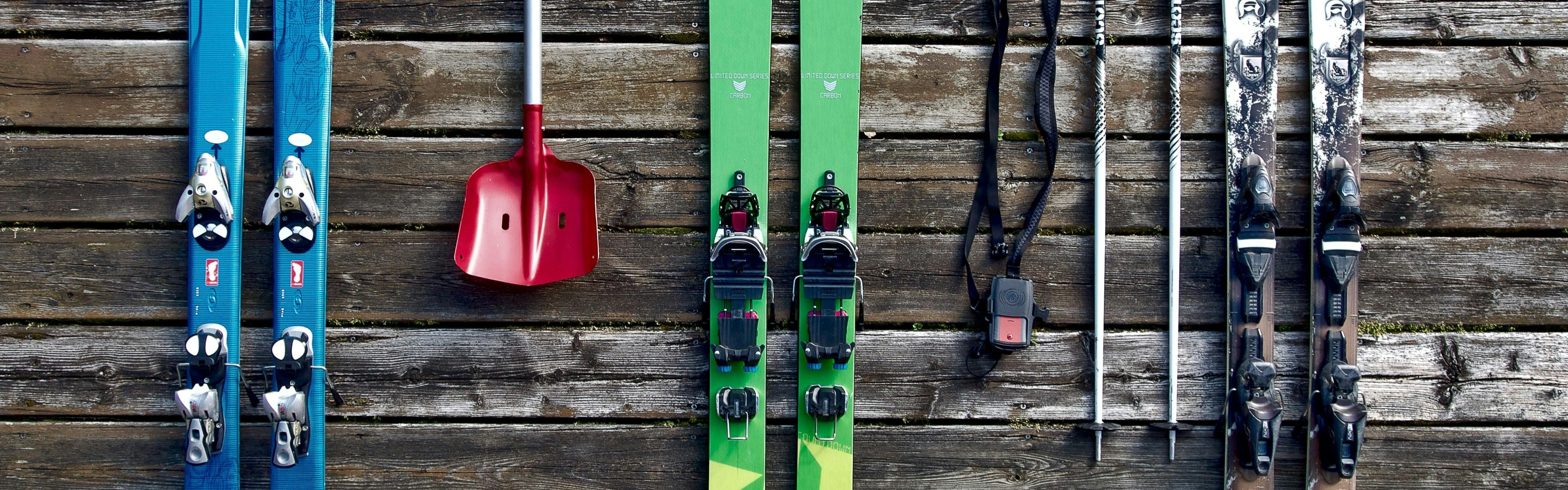 Three pairs of skis and a shovel laying on a wooden plank ground.