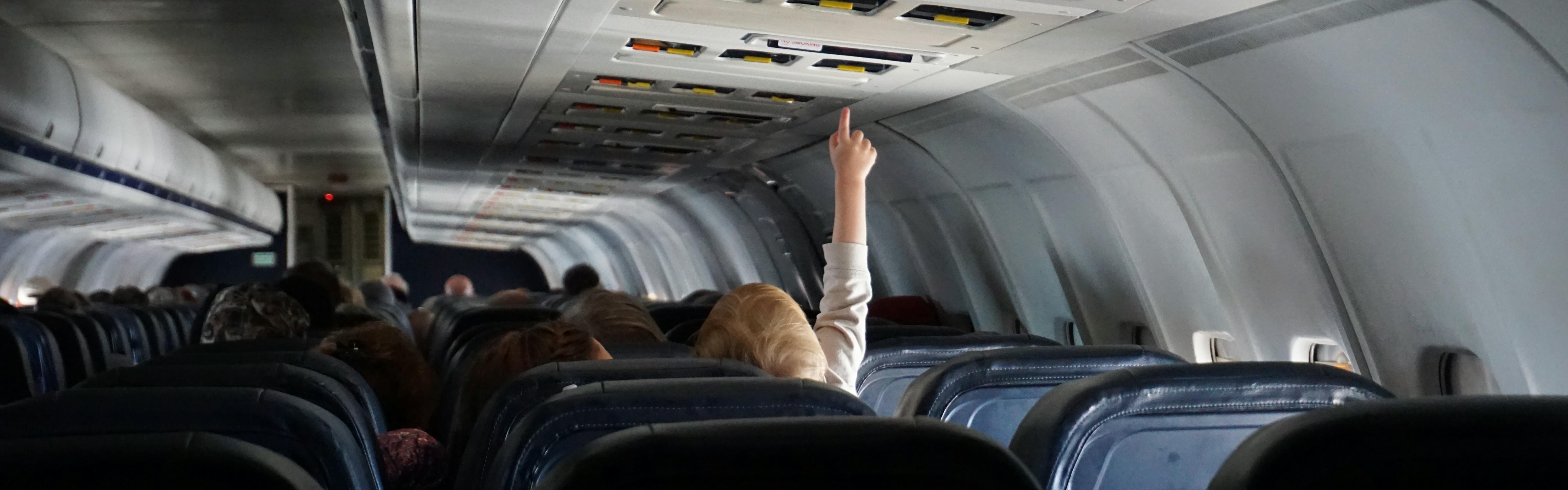 A baby reaches up from an airplane seat to touch a button on the ceiling. 