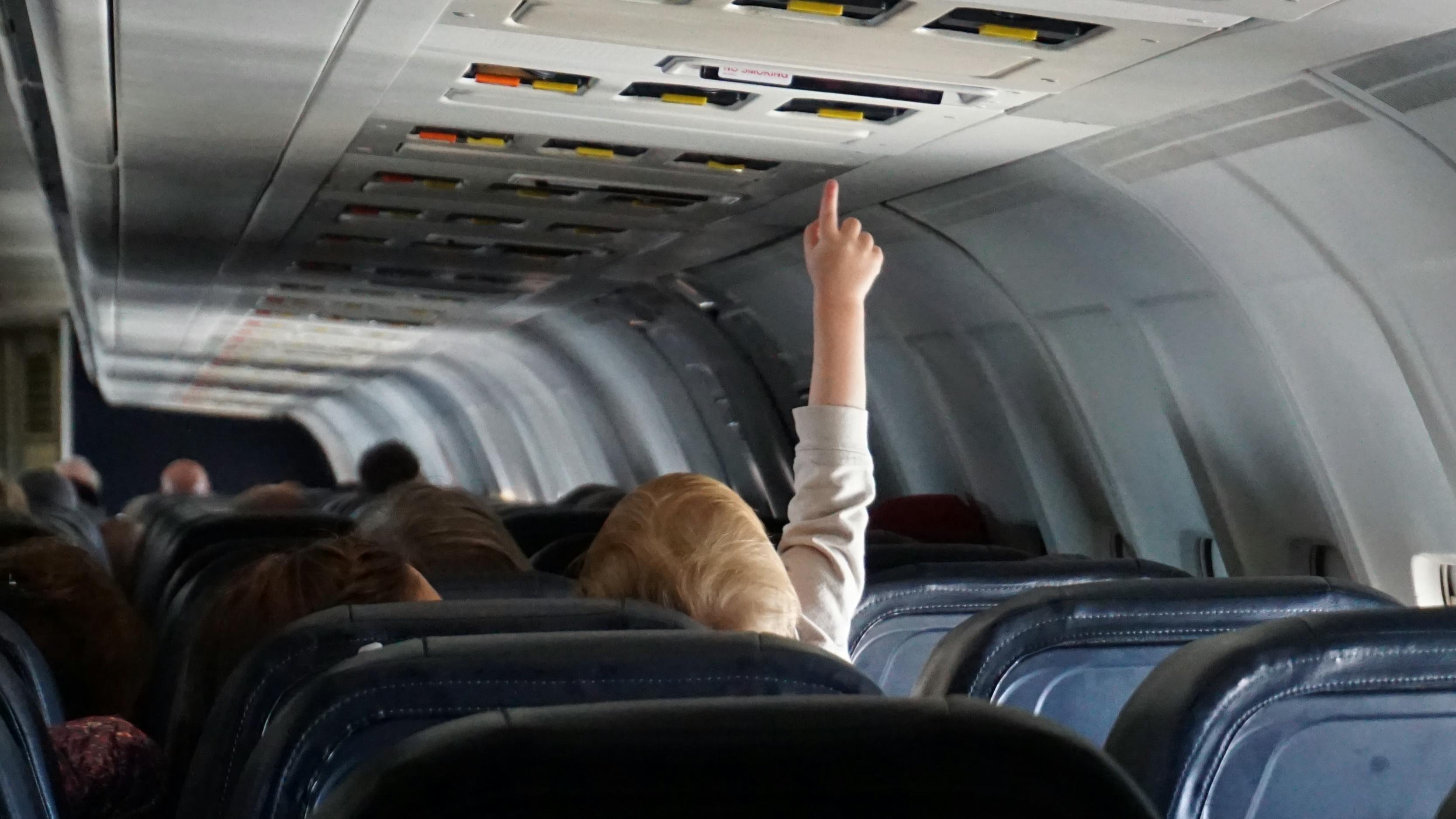 A baby reaches up from an airplane seat to touch a button on the ceiling. 