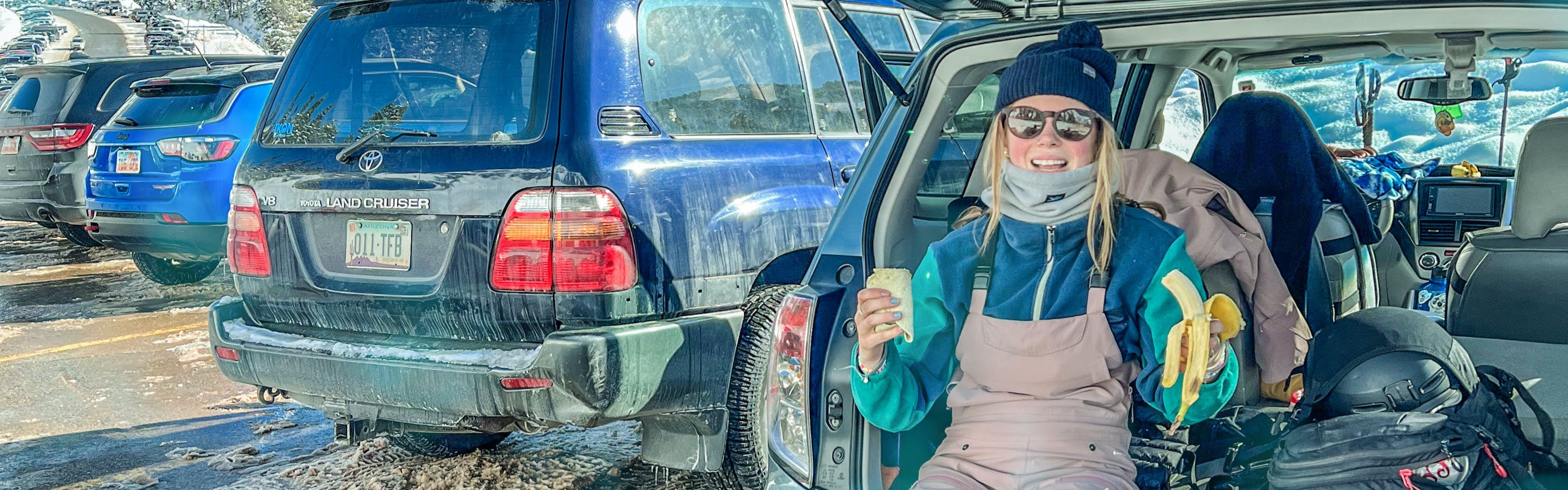 A skier sitting in the back of her car in a ski resort parking lot holding a burrito and a banana.