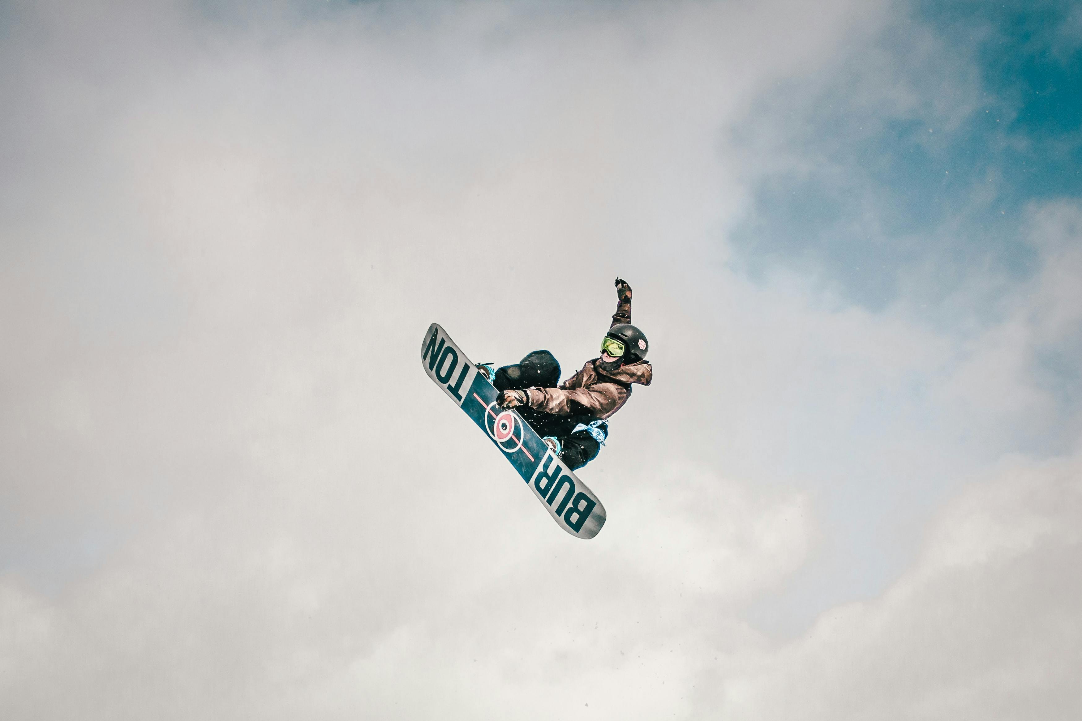 A shot from below of a snowboarder grabbing his snowboard while flying through the air. There is no ground in sight, only the man and his snowboard. He has one hand on the board and one up in the air waving. It is a little cloudy with some blue sky popping out. 