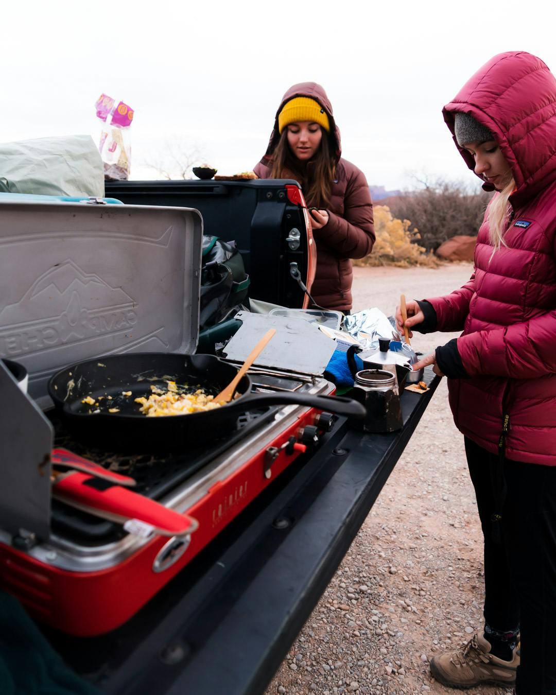 Two campers cooking on a tailgate with large, puffy jackets.