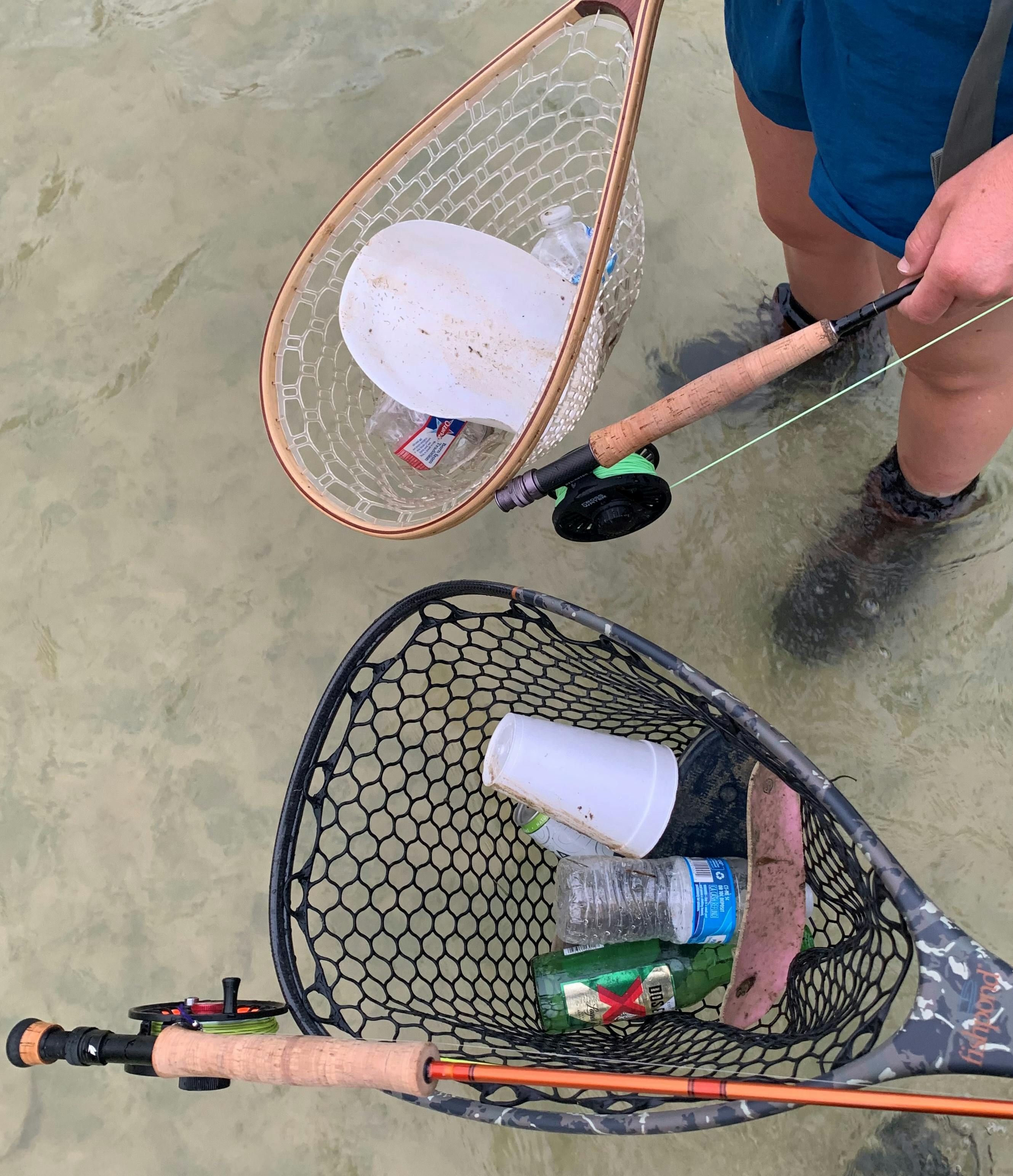 Two people carry nets filled with trash as they stand in the water. The image is taken from above, so we can see the trash in the nets, their rods in their hands, and their feet in the water.