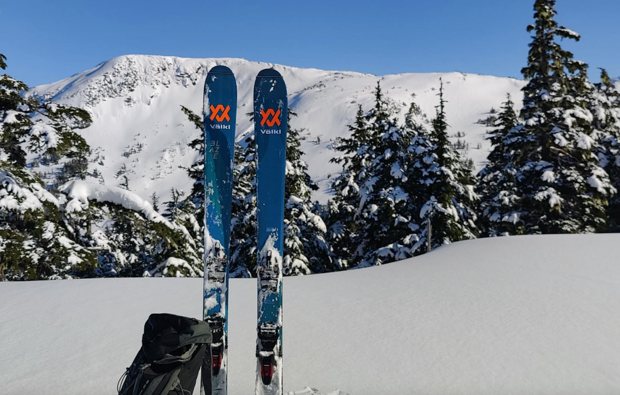 Two Volkl skis sticking out of the snow with a snowy mountain in the background. 