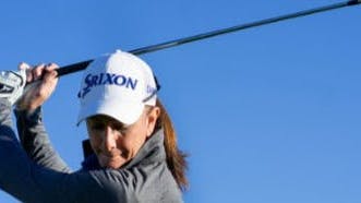 A woman wearing a hat swings a golf club.