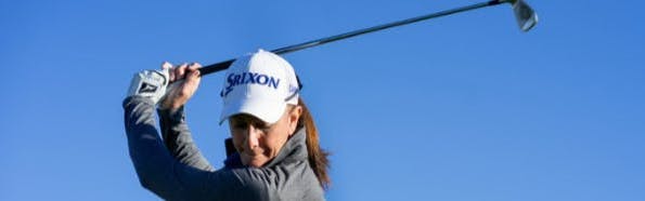 A woman wearing a hat swings a golf club.