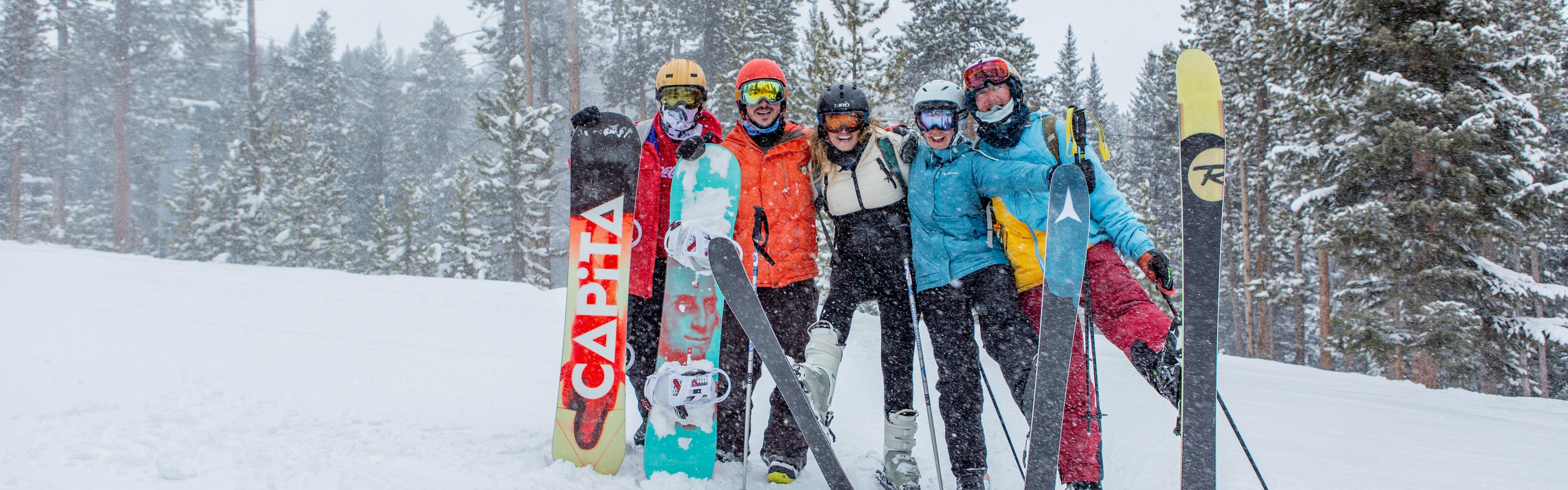 A group of three skiers and two snowboarders stand with their arms around each other and smile in the snow. 