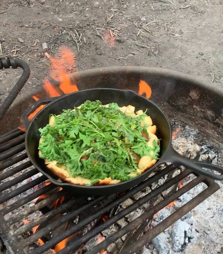 A pizza topped with a lot of greens sits in a cast-iron pan on a grill over a campfire. 