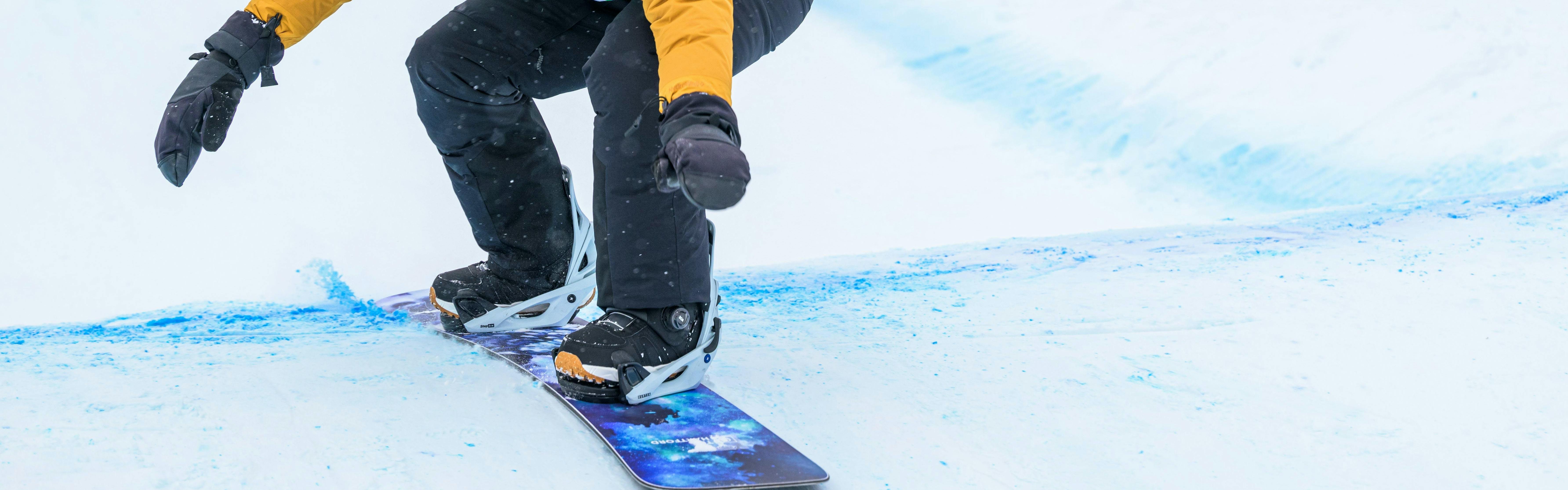 Snowboarder in yellow jacket and green race bib pumps through a roller section while racing on a banked slalom course.