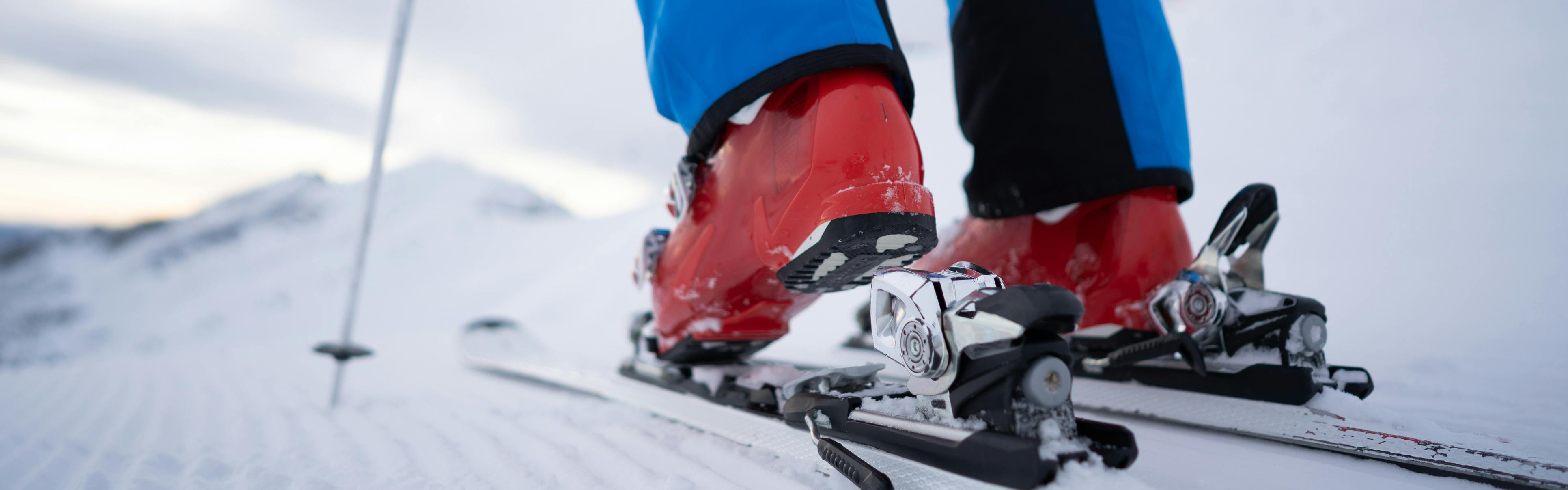 A person wearing red ski boots and blue pants steps into their skis