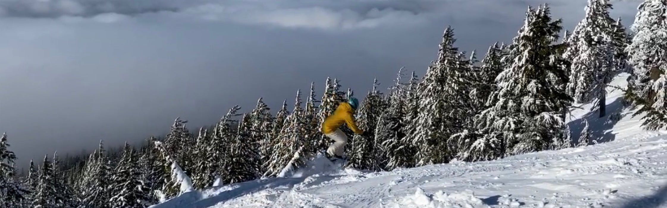 A snowboarder jumps off a small jump. There are trees and clouds in the background.
