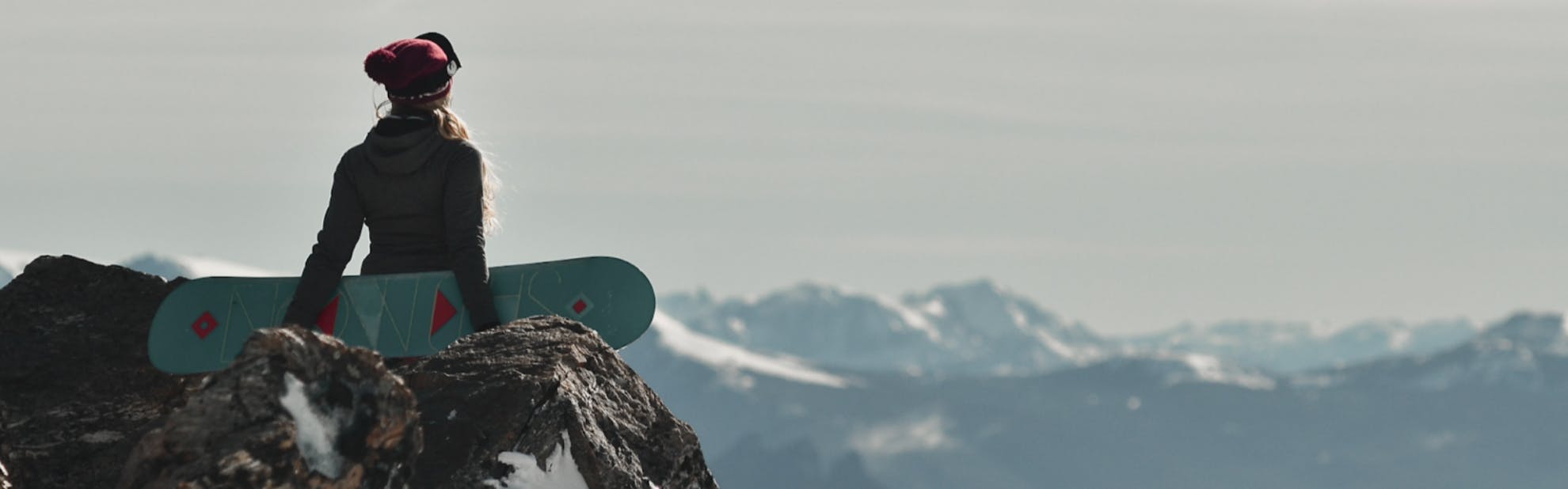 A woman standing looking out over a peak while holding her snowboard. 