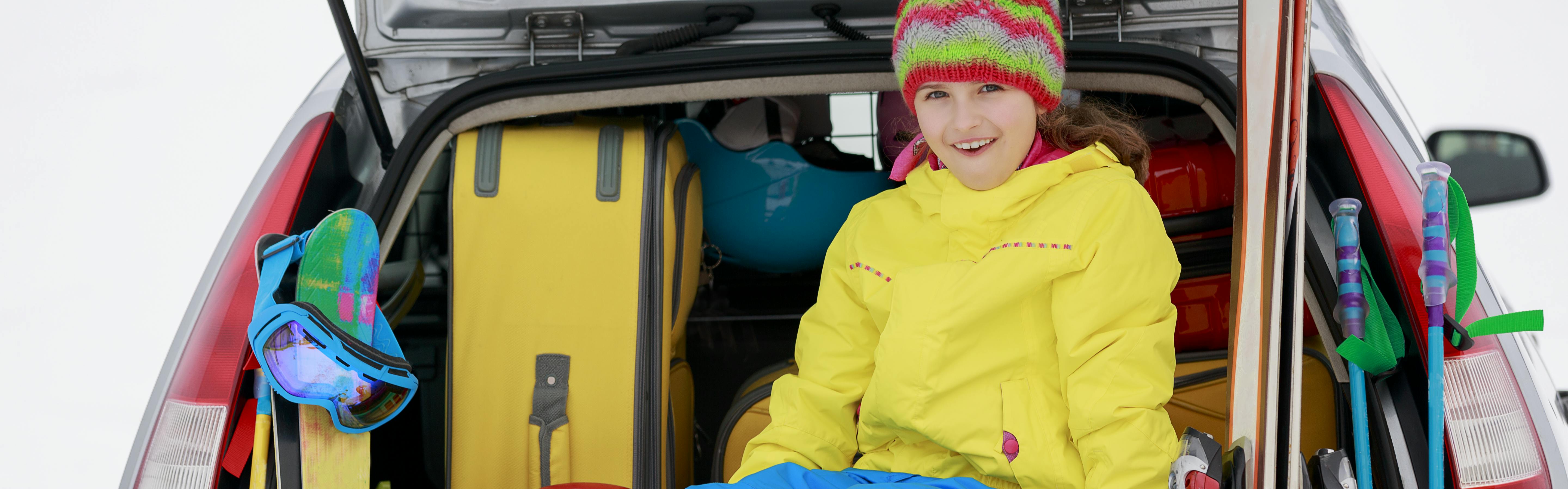 A young girl sitting in the trunk of a car smiling. There is a suitcase in the car, and ski gear surrounding the car. 