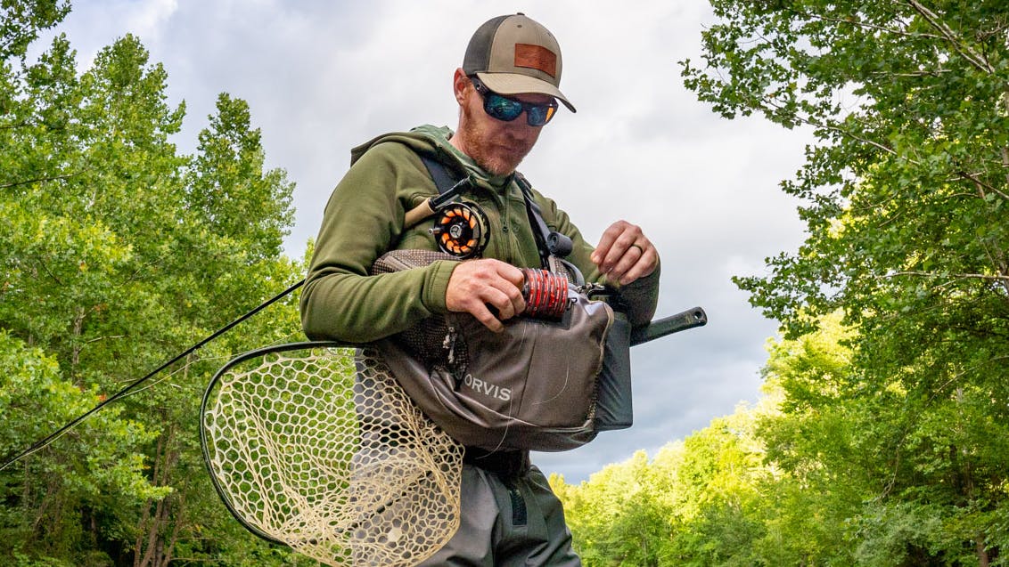 A man stands in a creek in waders and puts something into an Orvis bag while carrying his rod and a net. 