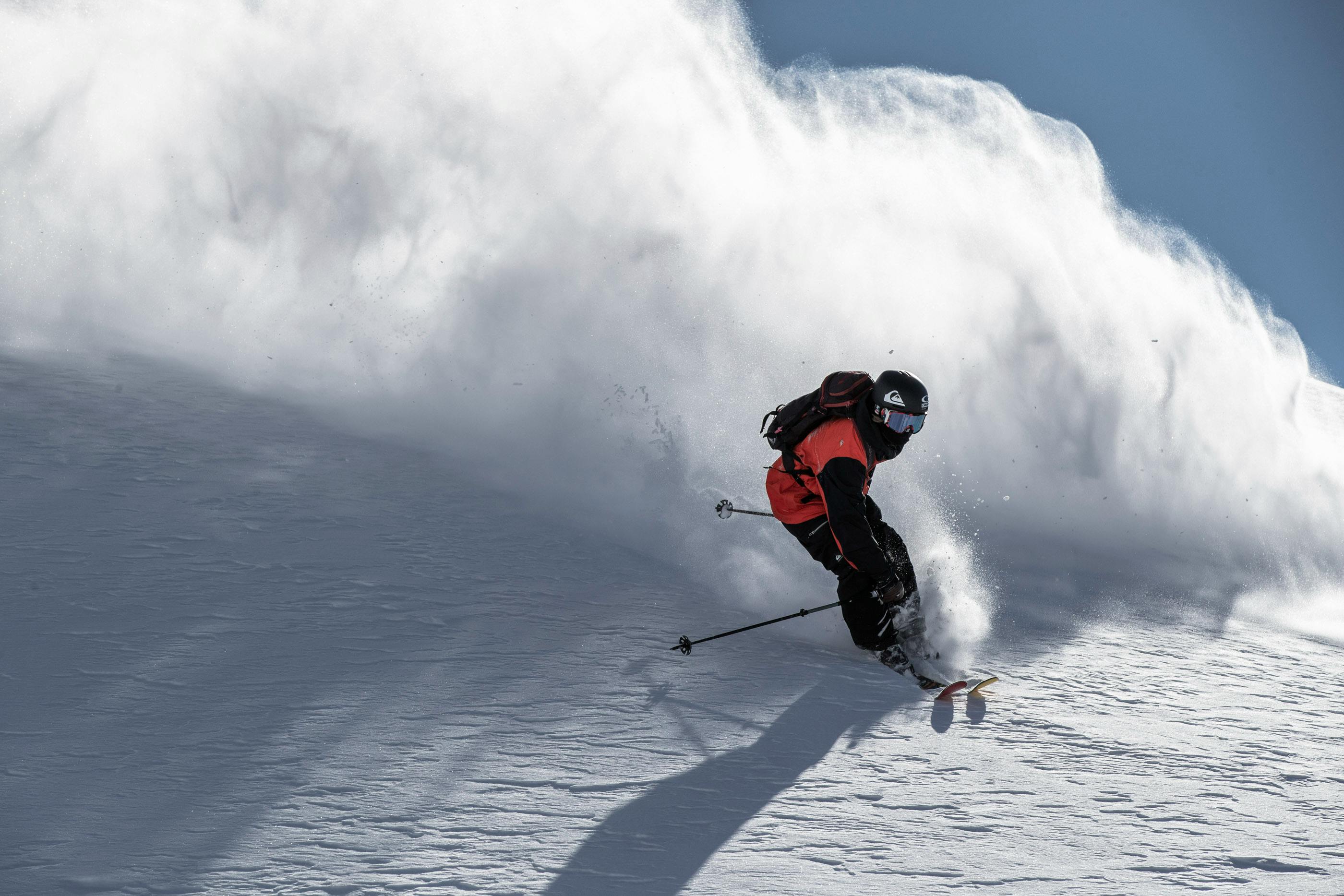 A male skier on Tour skis, holding his poles behind him, sending up a huge cloud of powder. 