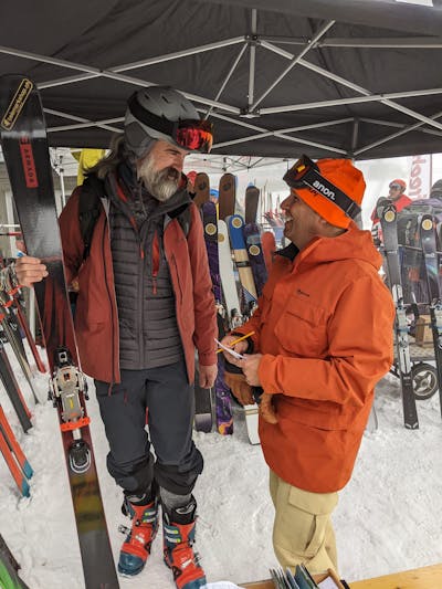 A man standing under a ski demo tent talking to another man. He is holding the Armada Declivity 88 skis.