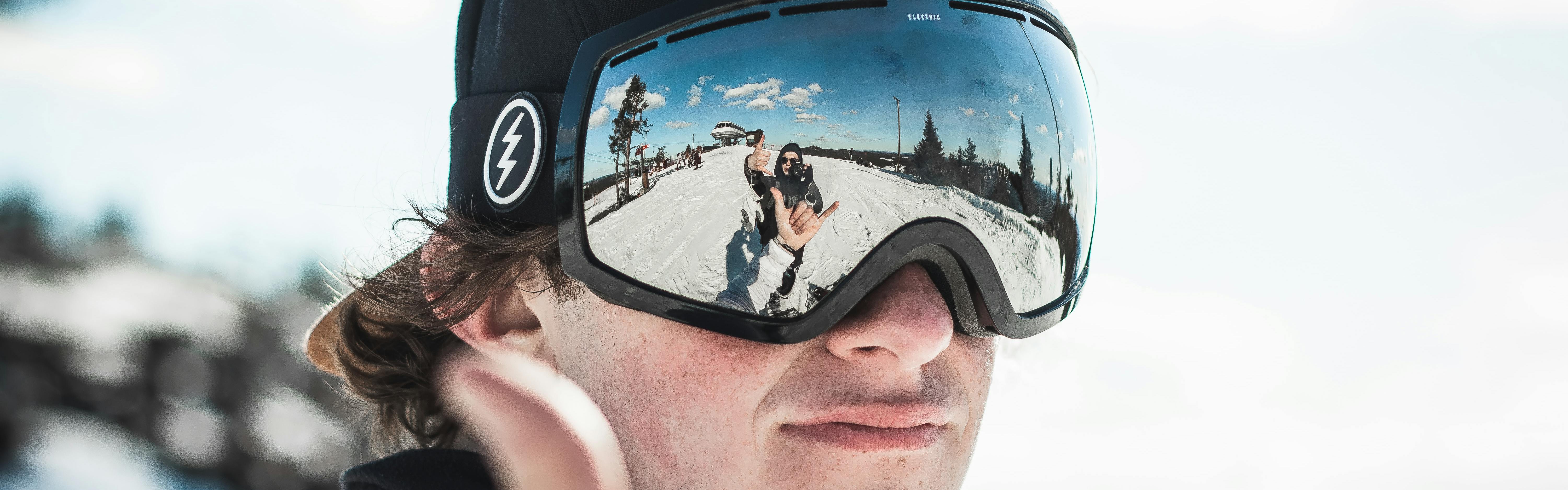 Close up of a snowboarders goggles. In the reflection of his goggles you can see his own hand and his friend waving with a camera. 
