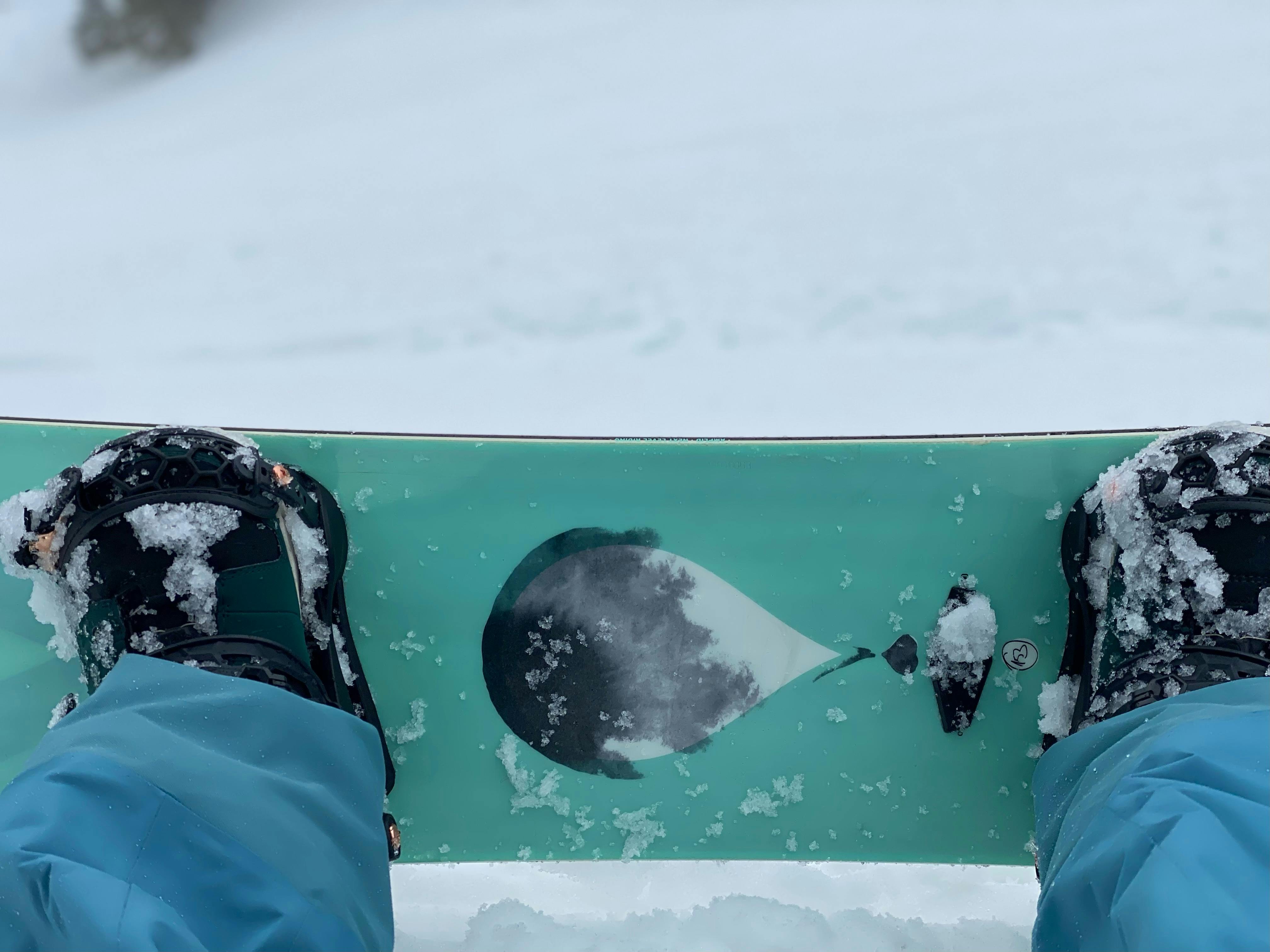 Top down view of a blue snowboard with black bindings. The bottom of the snowboarders pants are also blue and there is snow on the snowboard and underneath the snowboard.