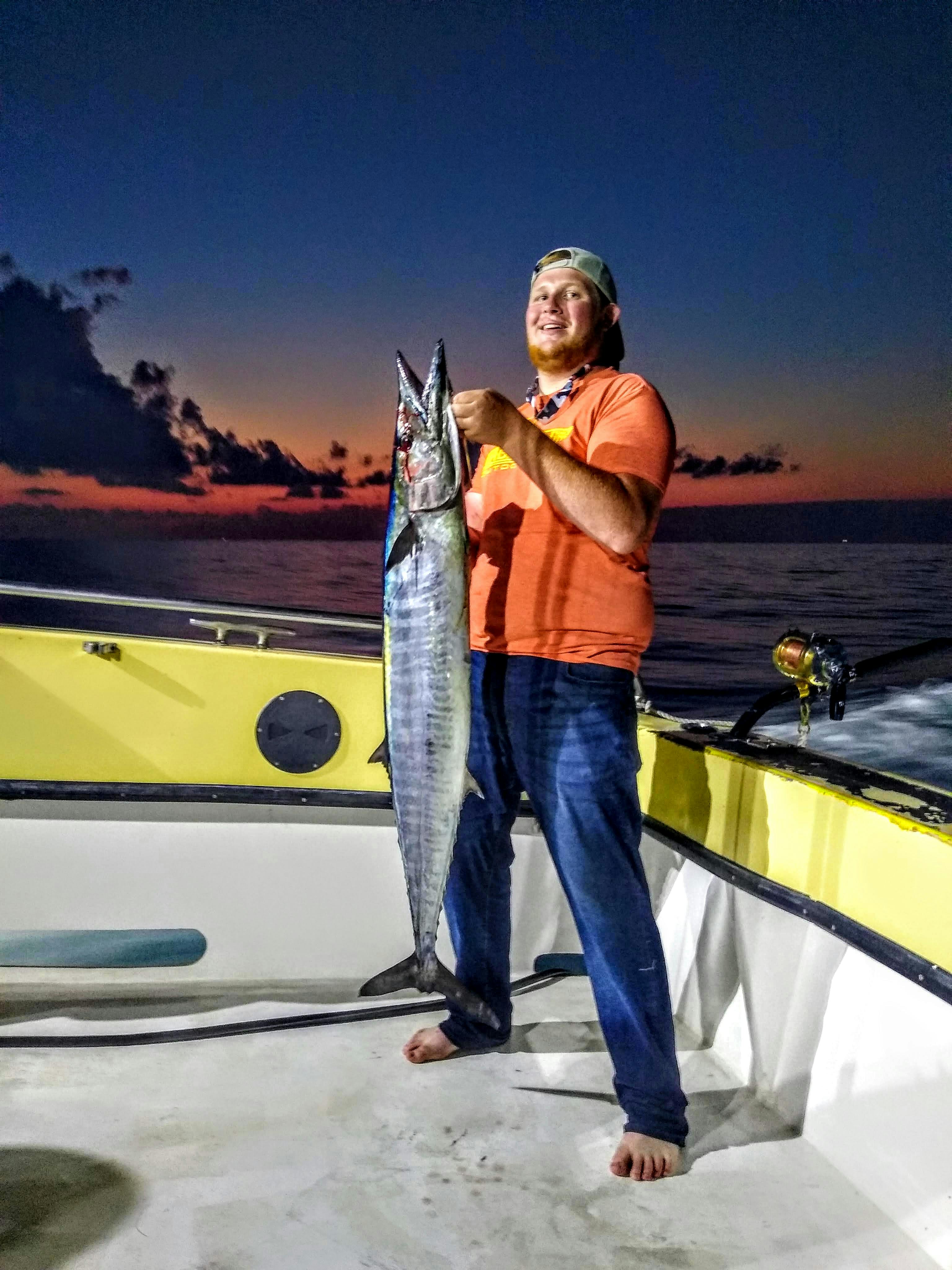 The author stands with bare feet on a boat on the open water. He holds a massive fish and smiles with it. The image is taken at twilight. 