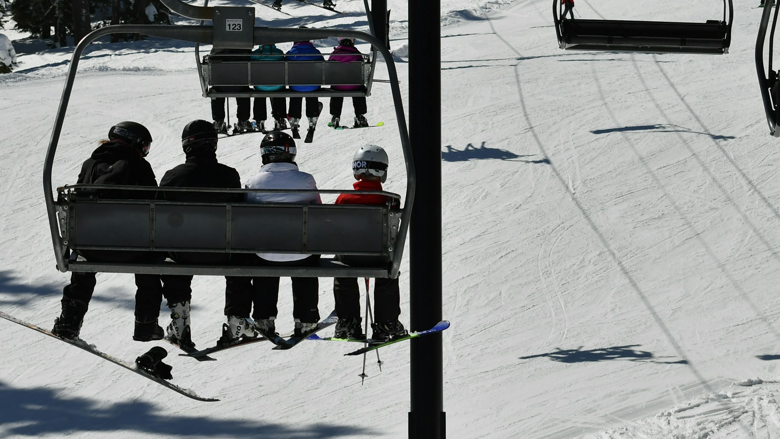 A family of 4 sits on a chairlift above a snowy slope. There is another 4-person group in front of them.