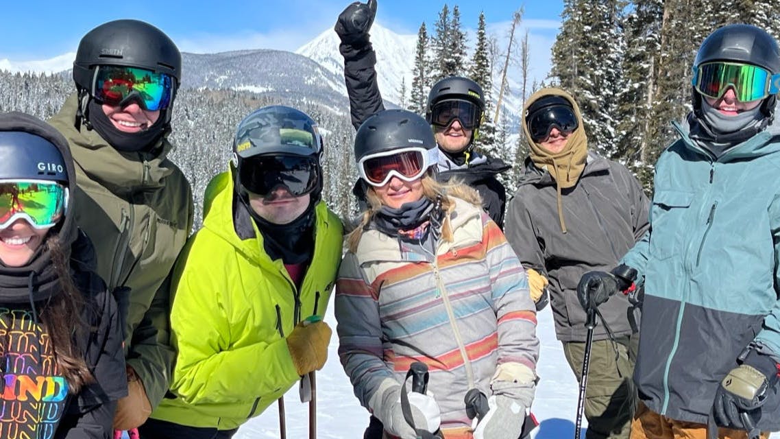 Several skiers and snowboarders posing together on a ski run. There are snowy trees in the background. 