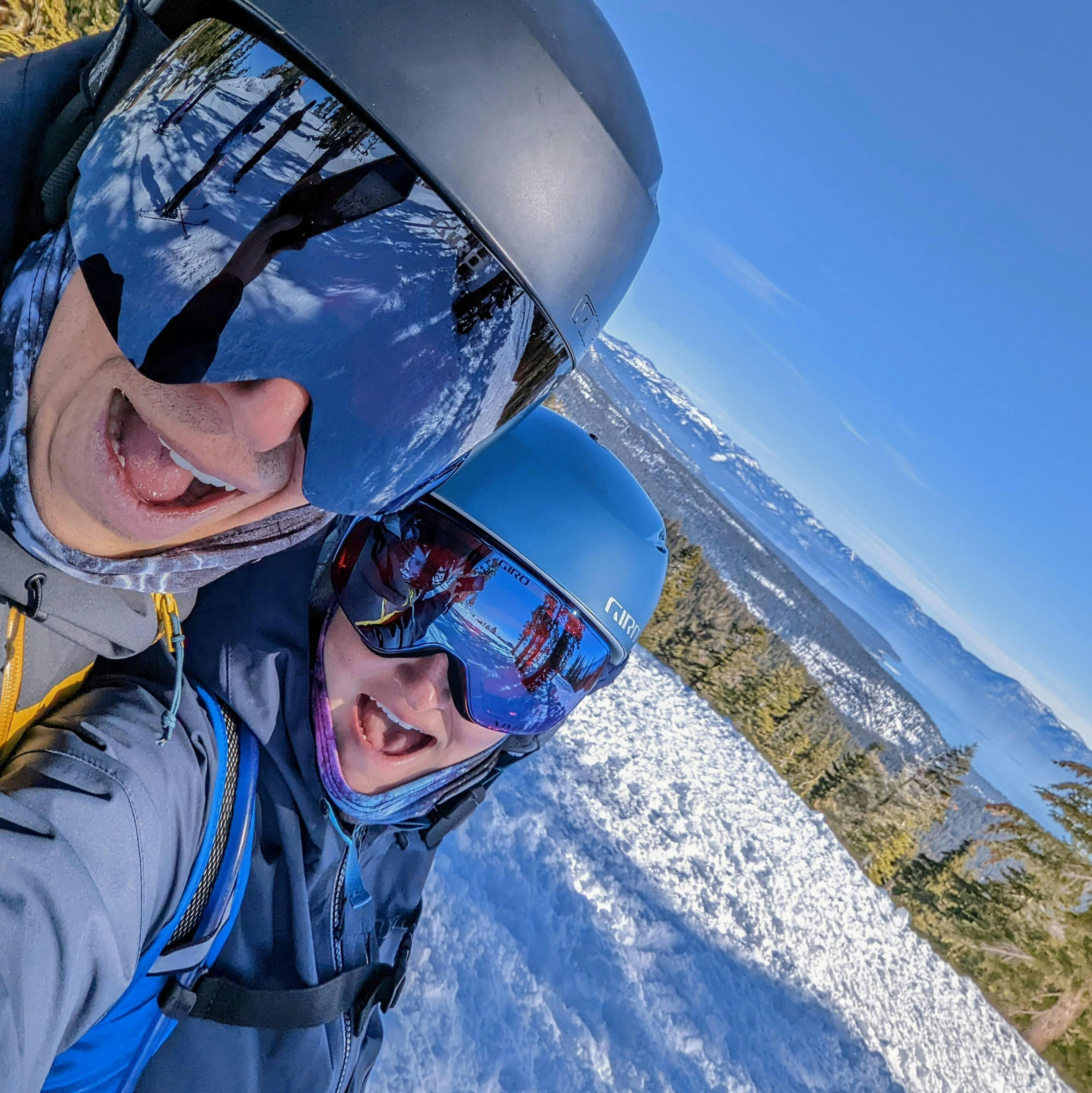 A selfie of two skiers with a snowy ski run in the background. 