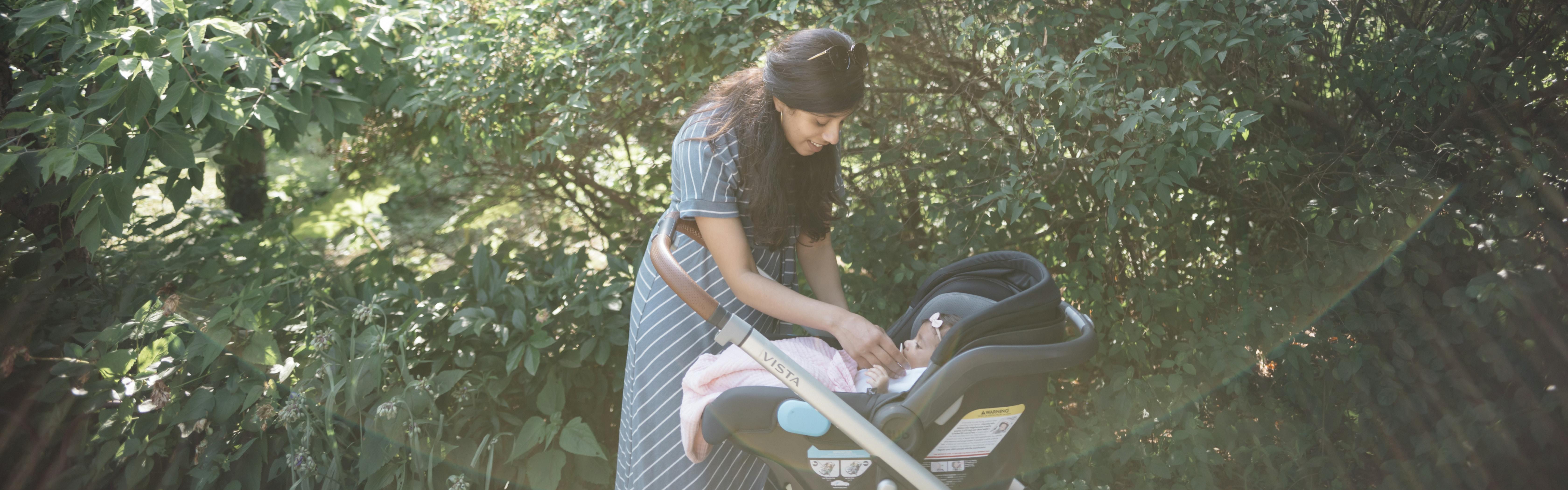 A woman smiles at a baby in a car seat. The car seat is in a stroller and the woman is standing near some foliage. 