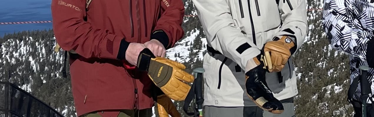Two people putting on their Hestra Freeride Czone 3 Finger Gloves with a ski resort in the background.