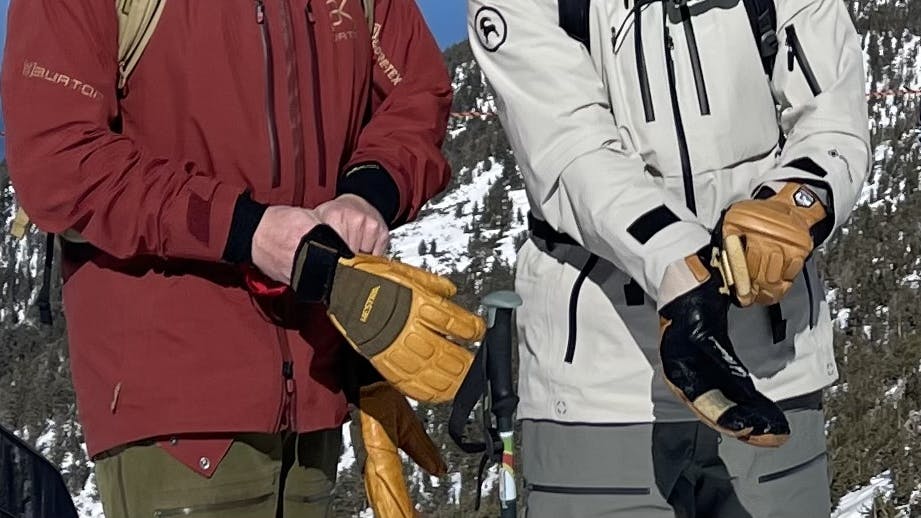 Two people putting on their Hestra Freeride Czone 3 Finger Gloves with a ski resort in the background.