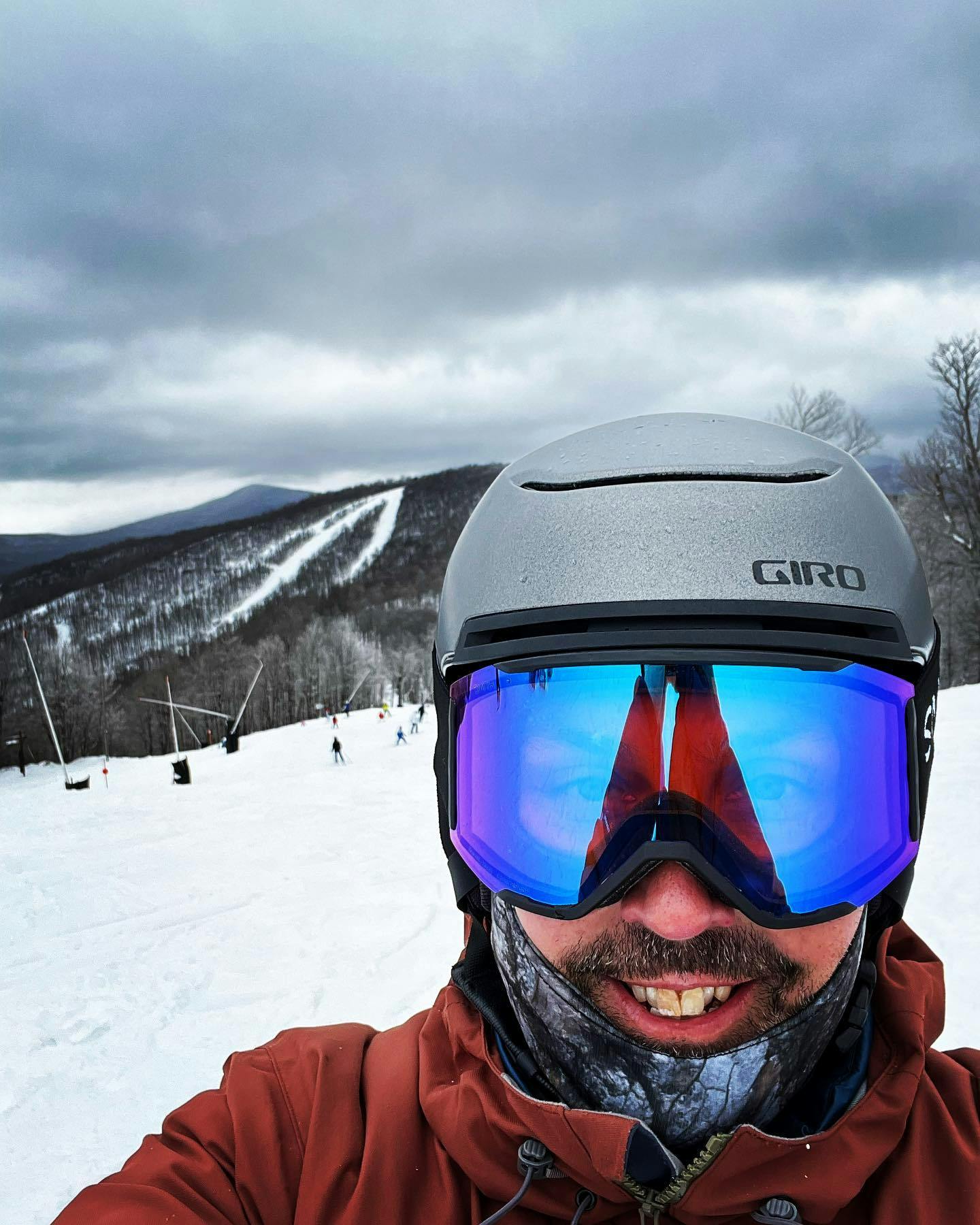 A skier taking a selfie while wearing a ski helmet and the Smith Squad MAG Goggles · 2022. There is a ski resort in the background. 