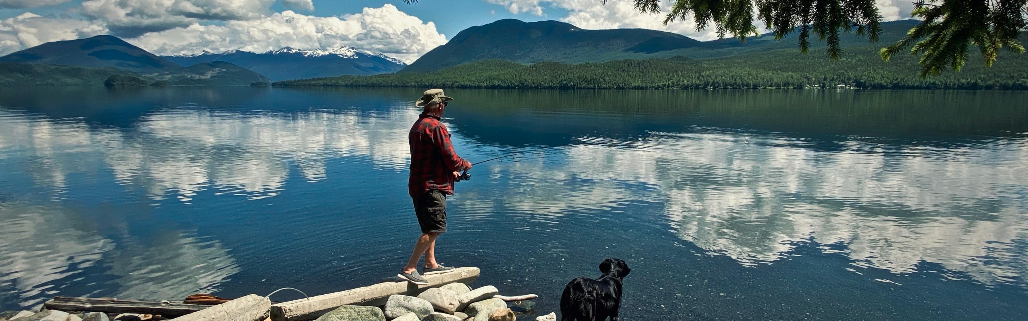 A man stands on rocks at a lake's edge and fishes. A black lab stands next to him.