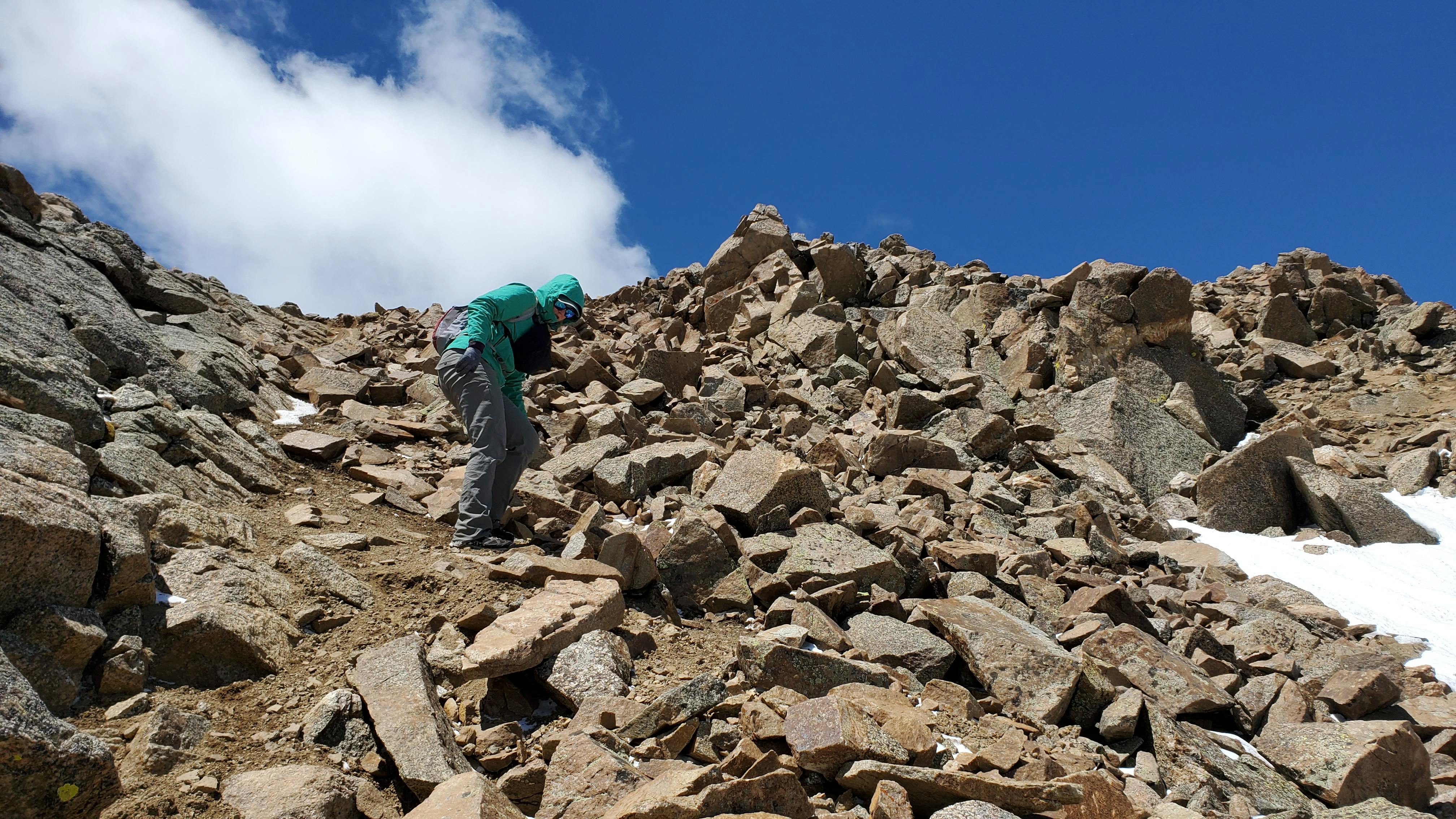 A hiker in a turquoise jacket and grey hiking pants descending from a rocky mountain summit