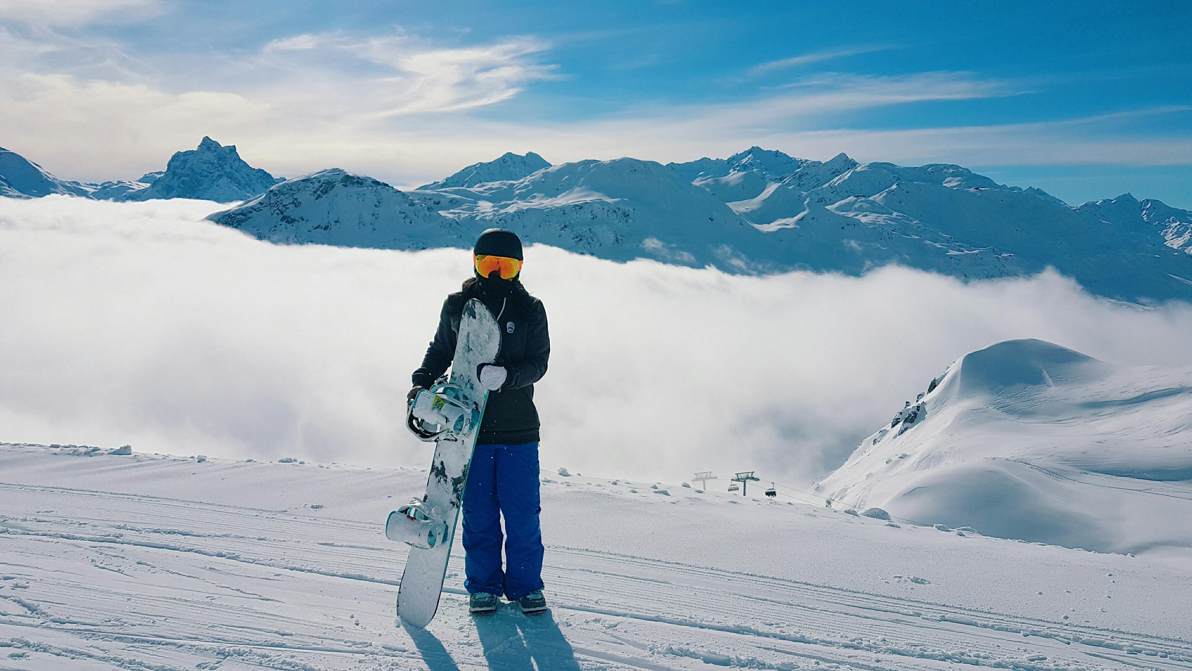 Person stands at the top of a mountain with a snowboard. There are clouds and mountain tops visible behind her.