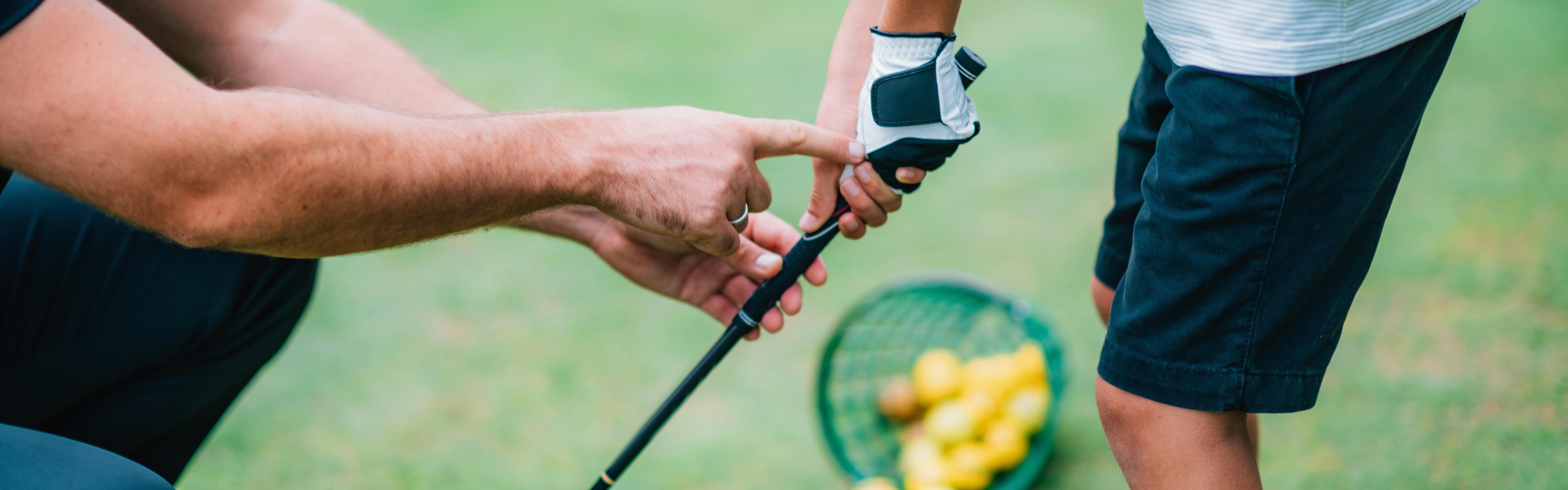 One set of hands teaching someone holding a golf club how to have proper hand placement. There is a bucket of golf balls in the background. 
