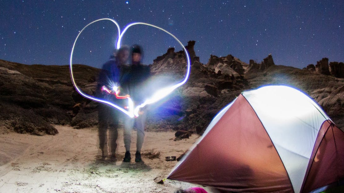 Two people standing together near a tent. There is a lit up heart around them. There is a desert landscape barely visible in the background.