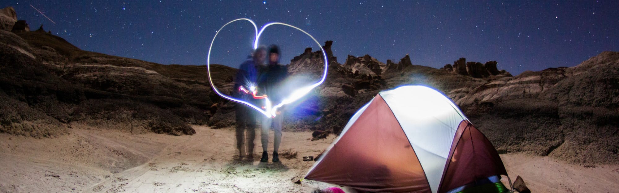 Two people standing together near a tent. There is a lit up heart around them. There is a desert landscape barely visible in the background.