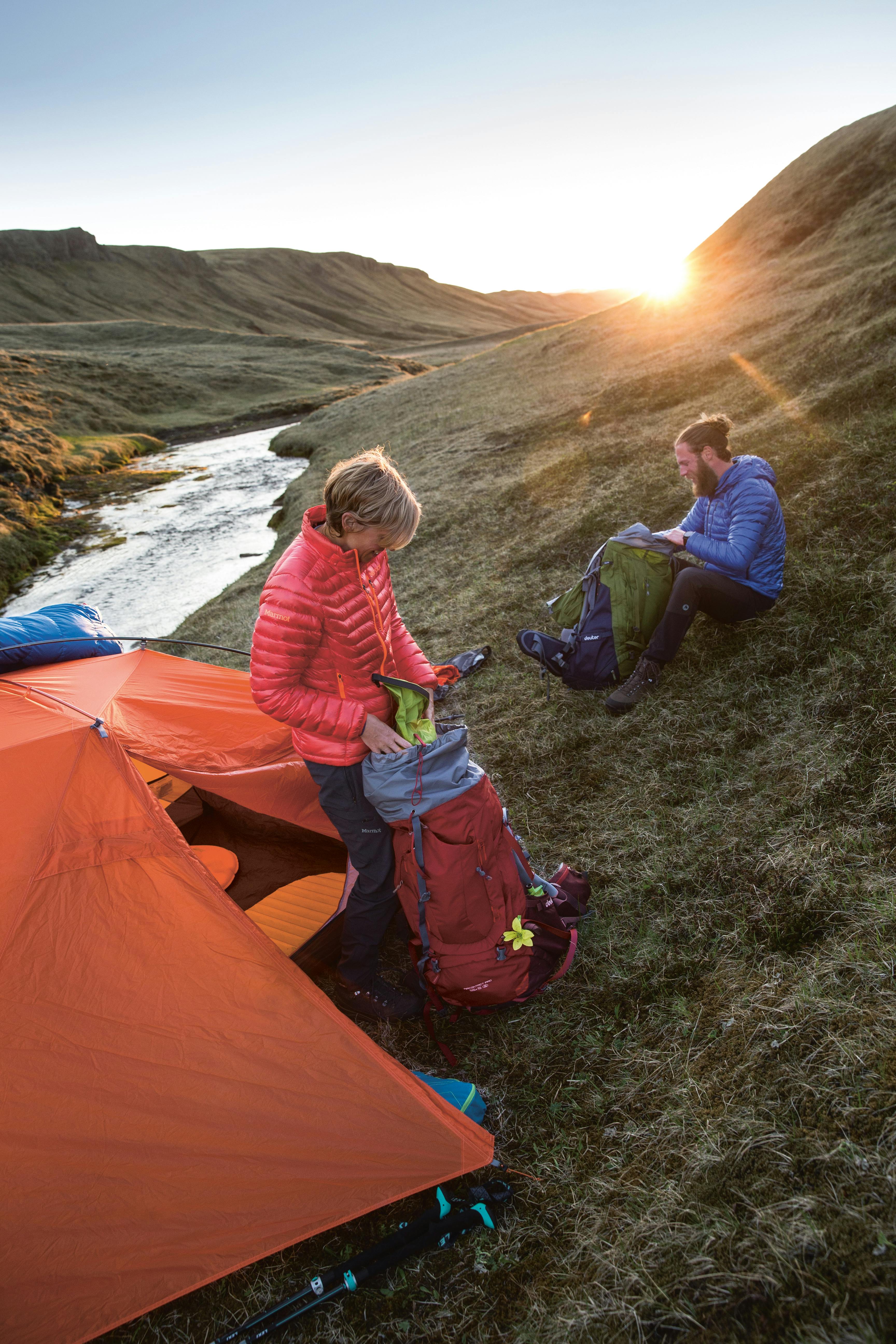 Two campers unloading their backpacks by an orange tent at their campsite by a stream winding through rolling grassy hills at sunset