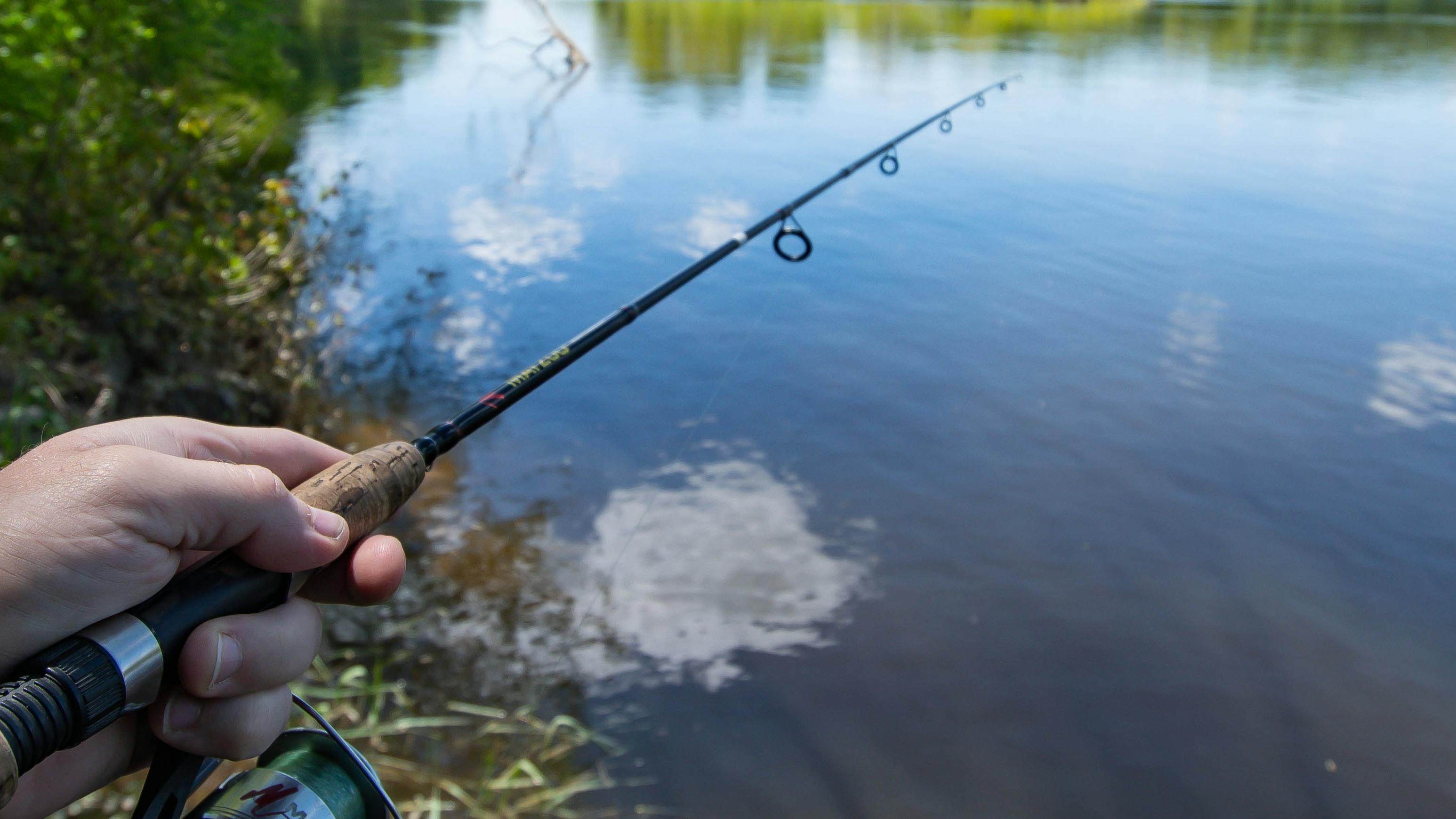 A hand on a conventional fishing rod. There is a body of water in the background. 
